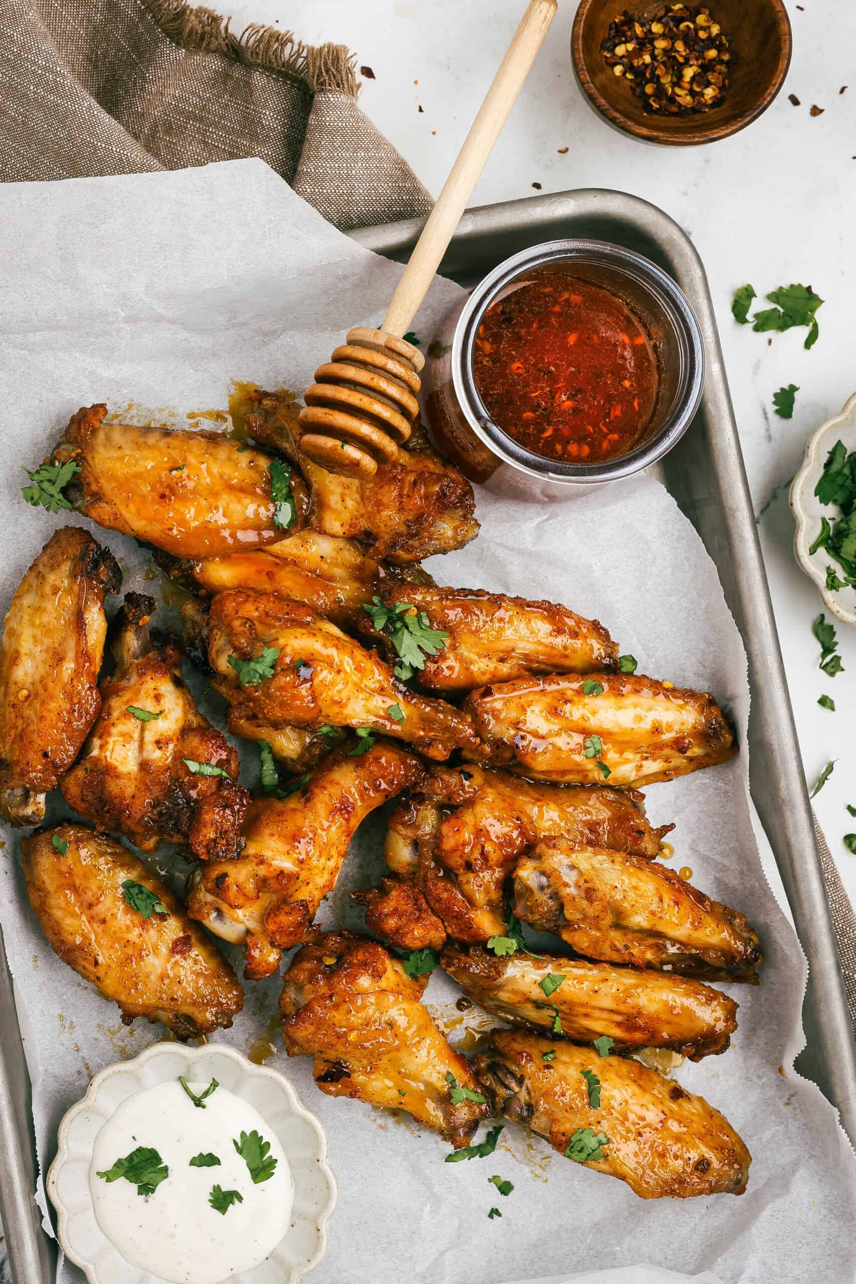 Golden oven-baked hot honey chicken wings on a parchment-lined tray with a honey dipper and bowl of hot honey glaze, garnished with cilantro.