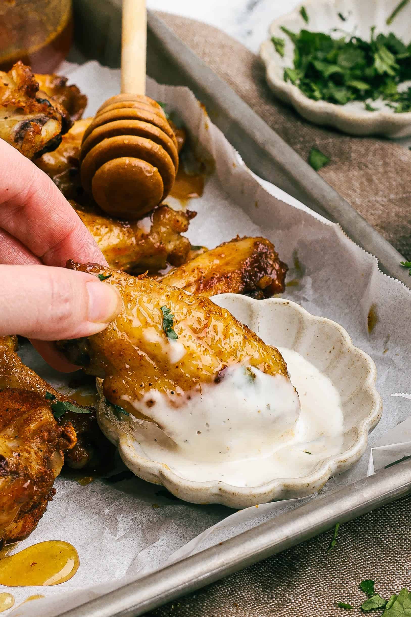 Close-up of a hand dipping a crispy hot honey chicken wing into a small dish of creamy ranch dressing, with honey drizzle and parsley garnish on a parchment-lined tray.