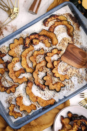 Maple roasted acorn squash slices spread across a parchment-lined baking sheet, surrounded by warm gold linens, rosemary, and kitchen utensils.