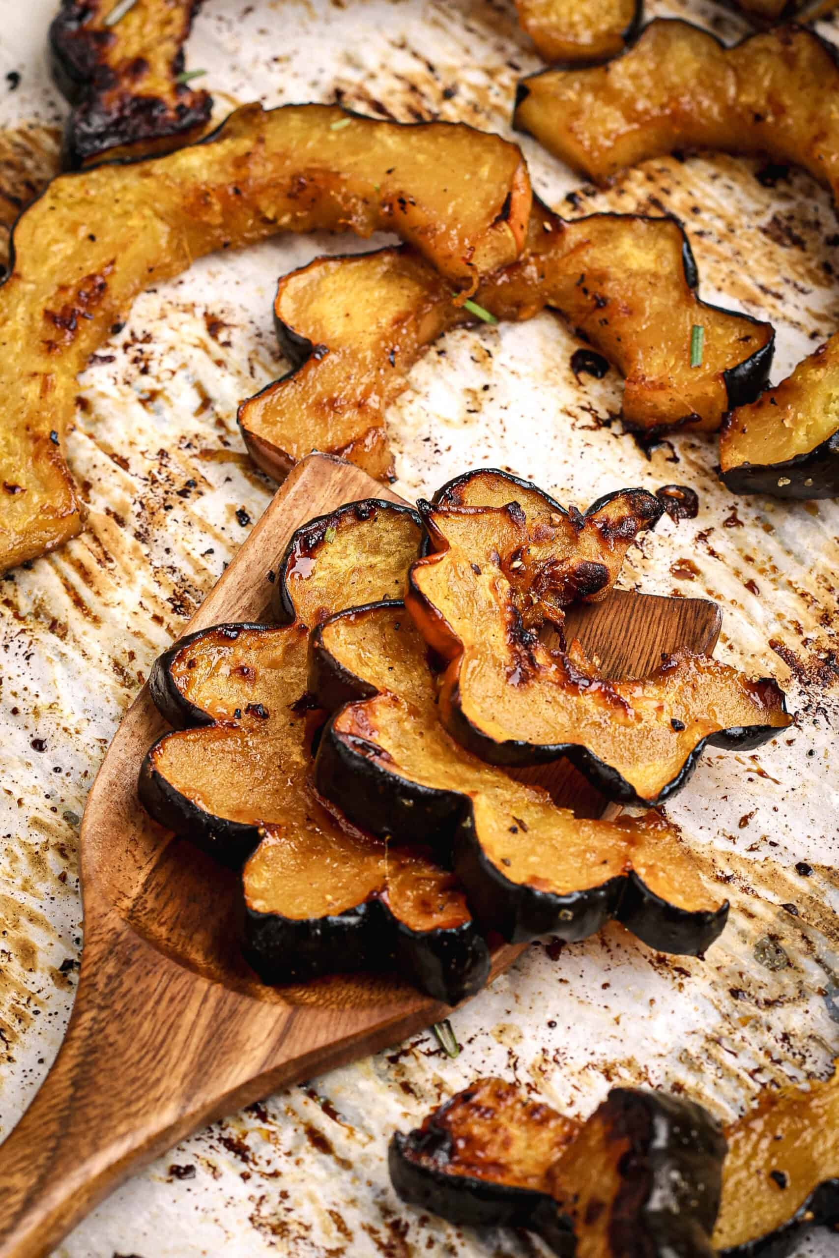 Roasted acorn squash slices resting on a wooden spatula over a parchment-lined baking sheet, showing caramelized edges and a rustic texture.