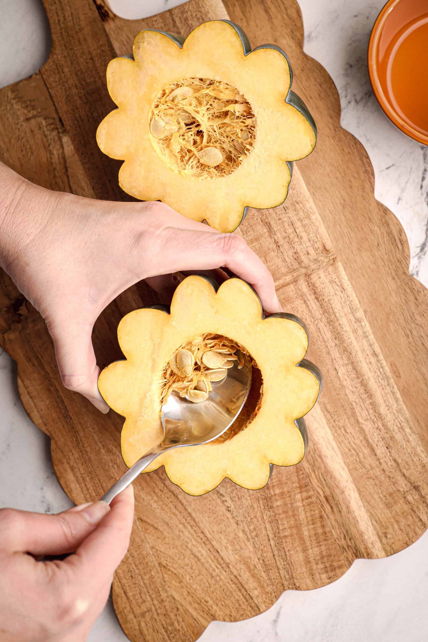 Hands using a spoon to scoop out seeds and pulp from a halved acorn squash on a wooden cutting board.
