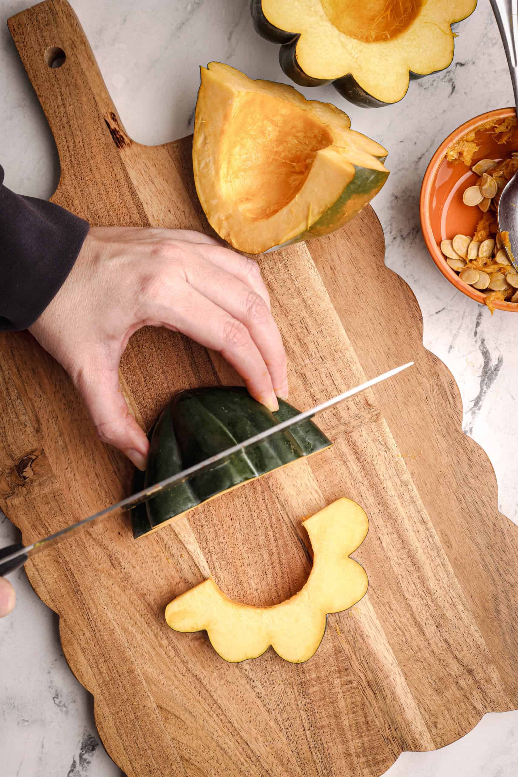 A hand holds a sliced acorn squash half as a knife cuts it into crescent-shaped pieces.