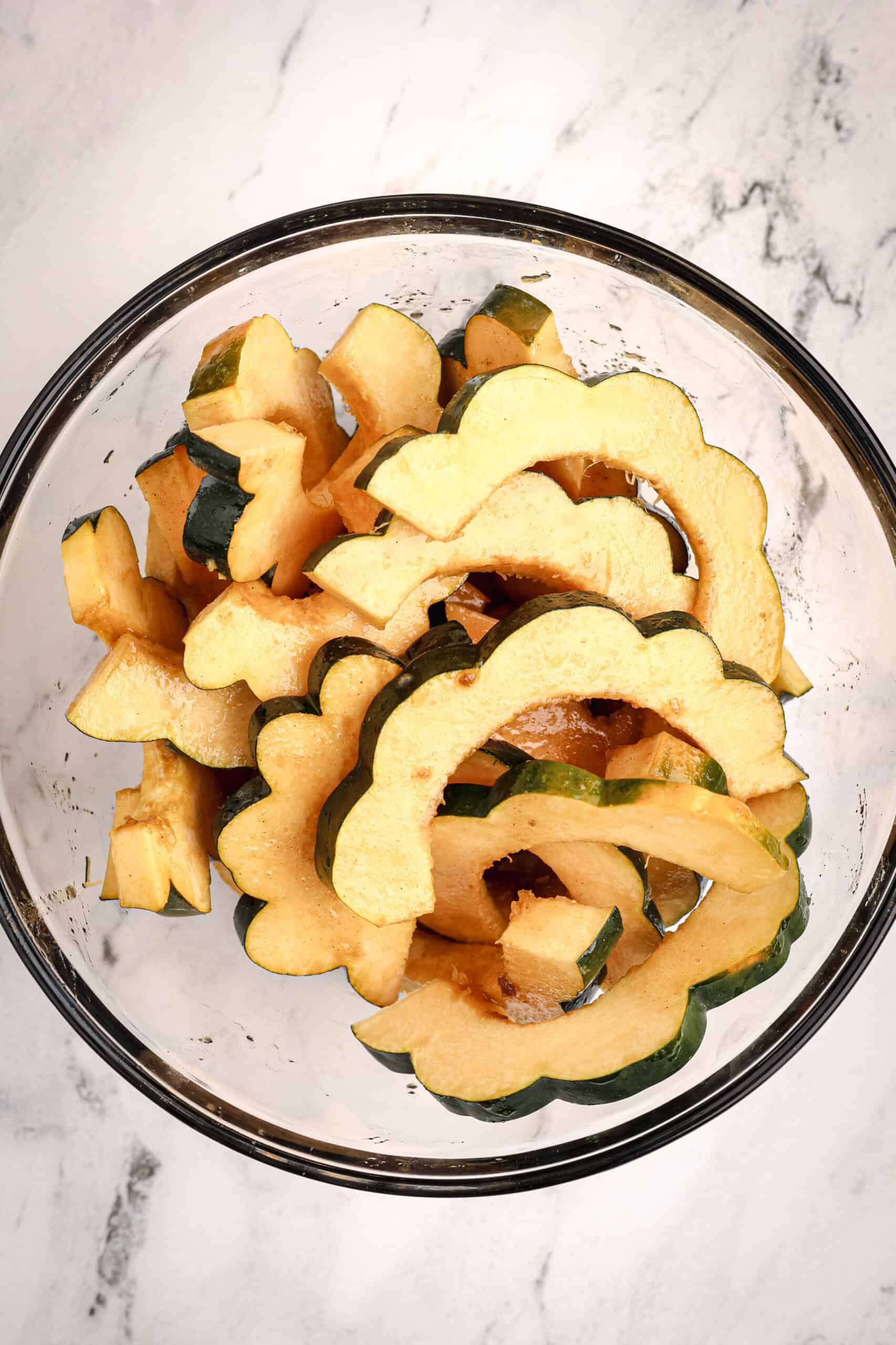 A glass bowl filled with raw acorn squash slices coated in maple glaze, ready for roasting.