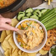 Bowl of buffalo chicken dip surrounded by tortilla chips, sliced cucumbers, celery sticks, and pretzel crisps, with a wooden spoon scooping into the dip.