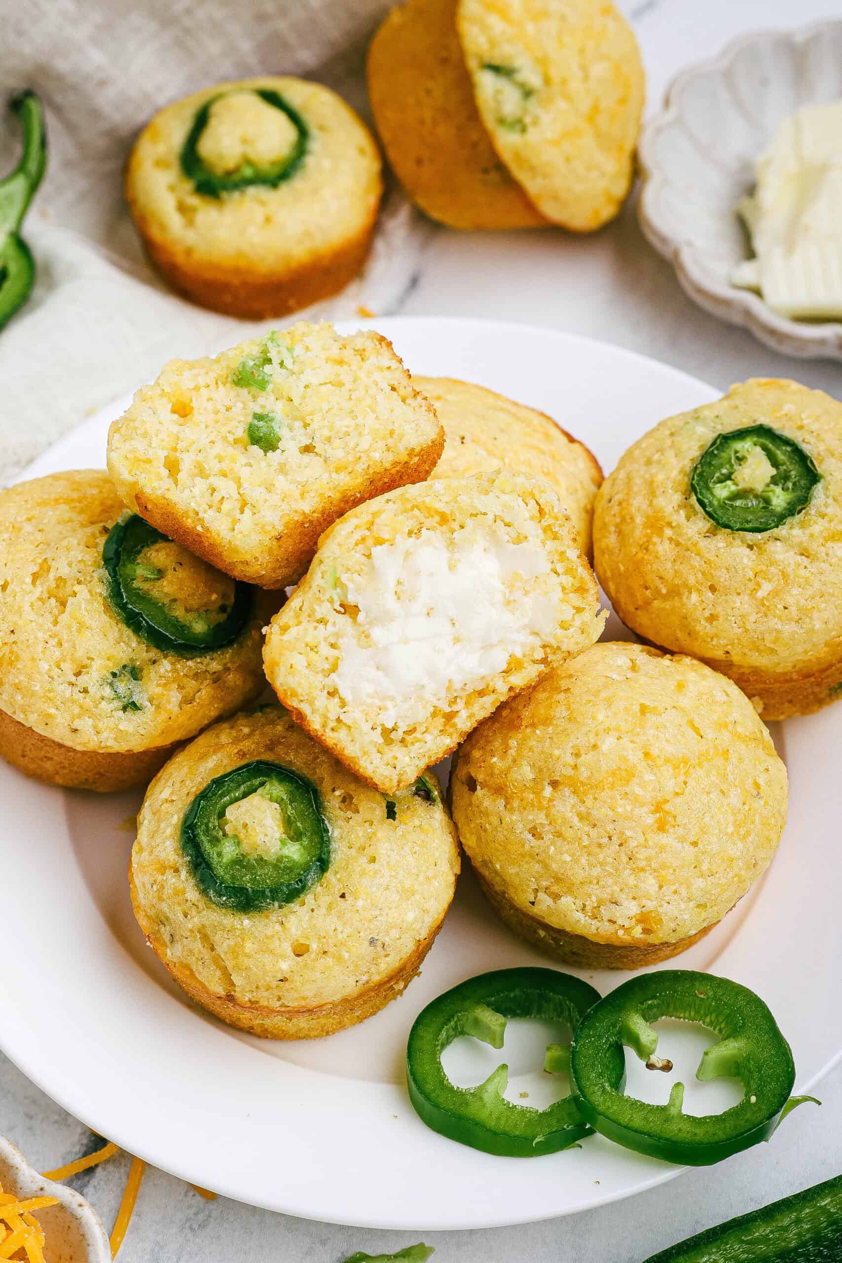Plate of mini jalapeño cheddar cornbread muffins with one muffin cut open and spread with butter, showing the fluffy interior and bits of green jalapeño.