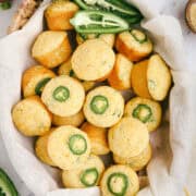 Basket lined with linen cloth filled with freshly baked mini jalapeño cheddar cornbread muffins, each topped with a jalapeño slice, surrounded by shredded cheese and fresh peppers.