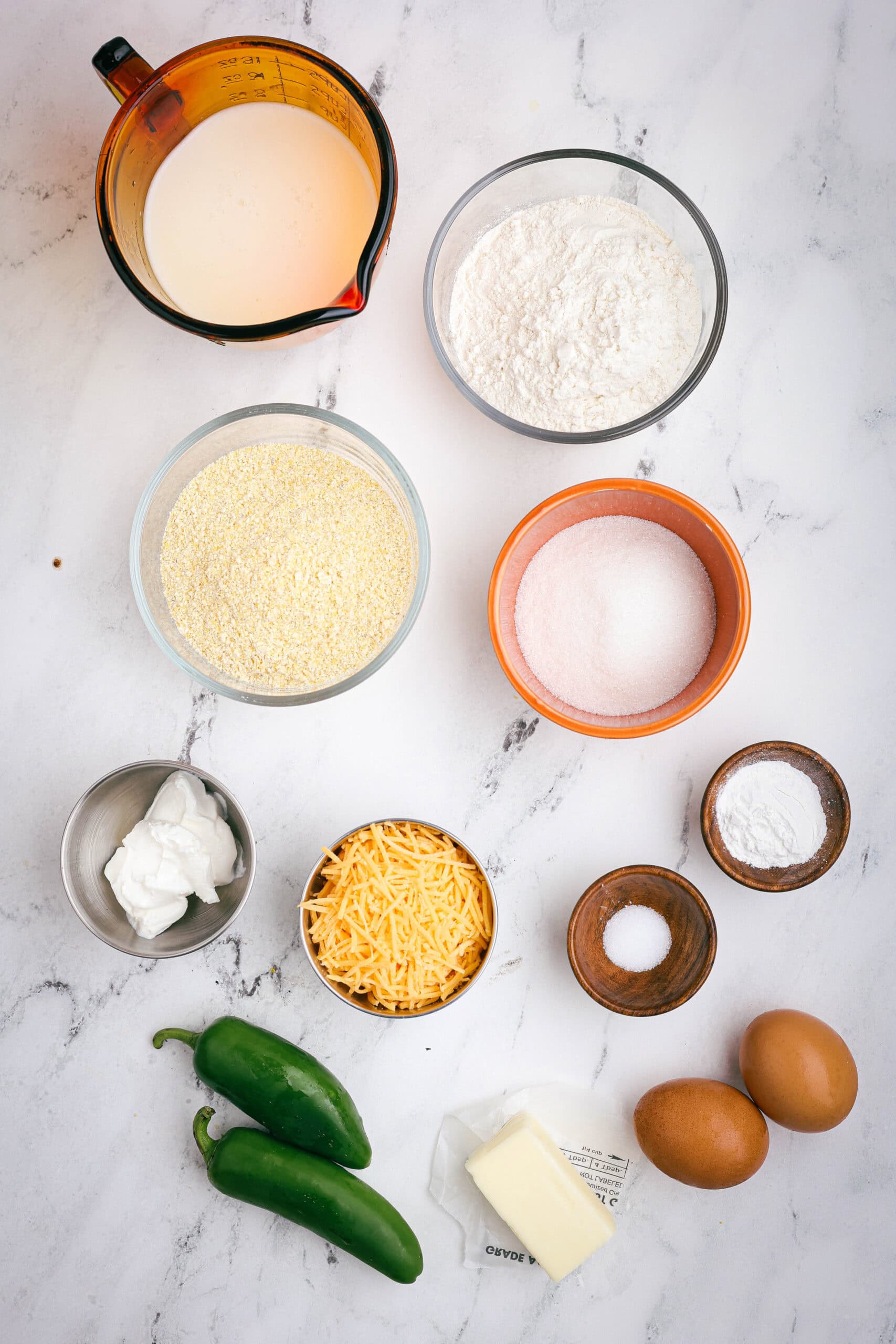 Flat lay of ingredients for mini jalapeño cheddar cornbread muffins including cornmeal, flour, sugar, eggs, butter, sour cream, cheddar cheese, and fresh jalapeños on a marble surface.