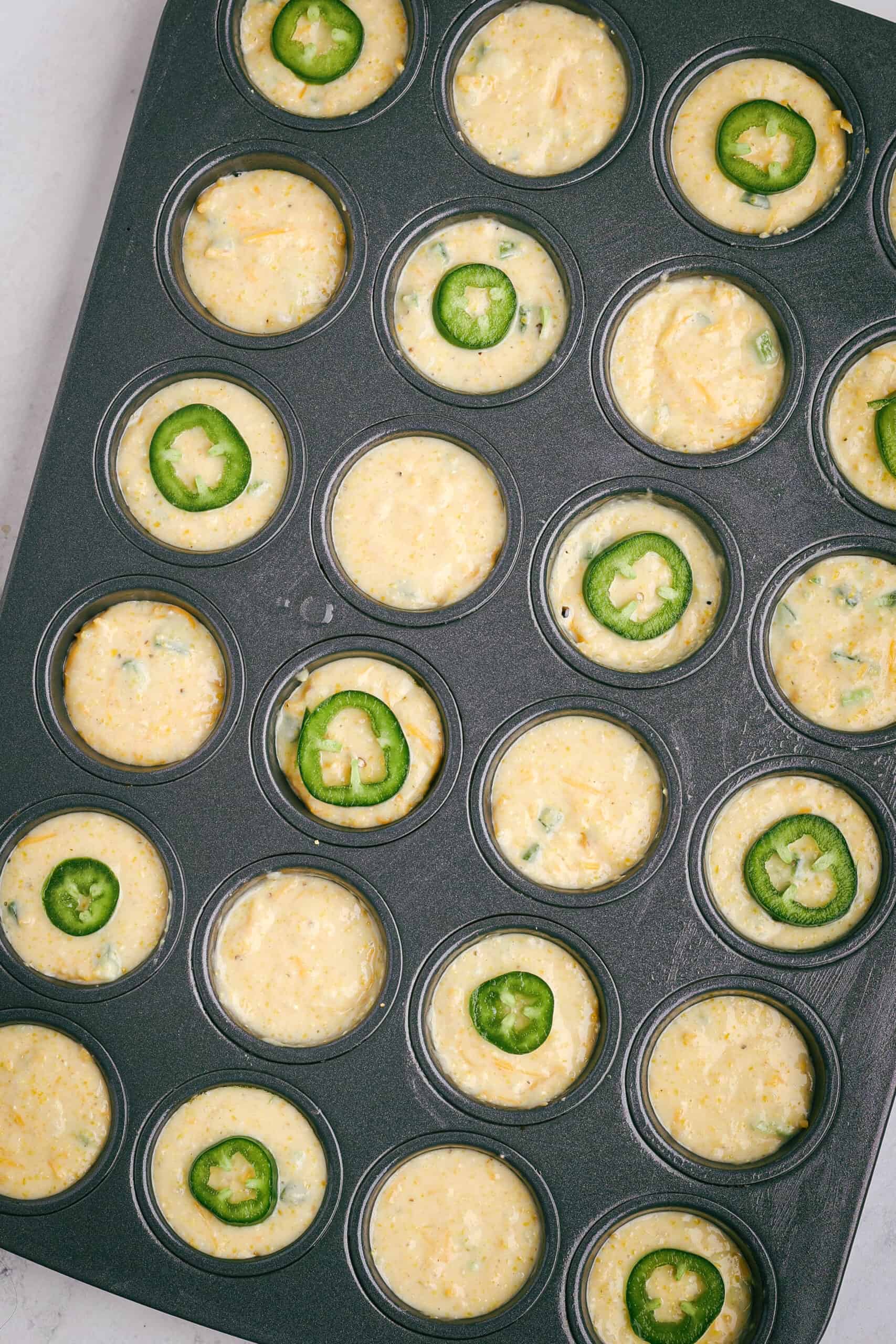 Mini muffin tin filled with cornbread batter, each topped with a slice of jalapeño before baking.