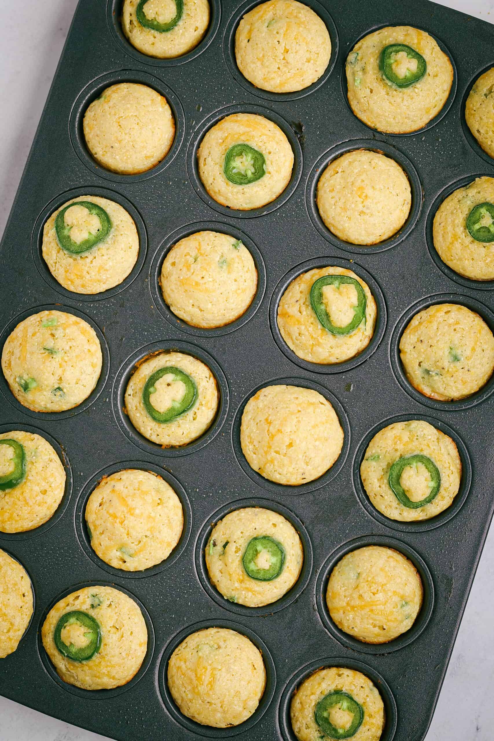 Baked mini jalapeño cheddar cornbread muffins still in the tin, golden brown and puffed with crisp edges and green jalapeño slices on top.