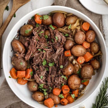 Shredded slow cooker pot roast served in a large white bowl with carrots, red potatoes, and onions, garnished with fresh herbs.