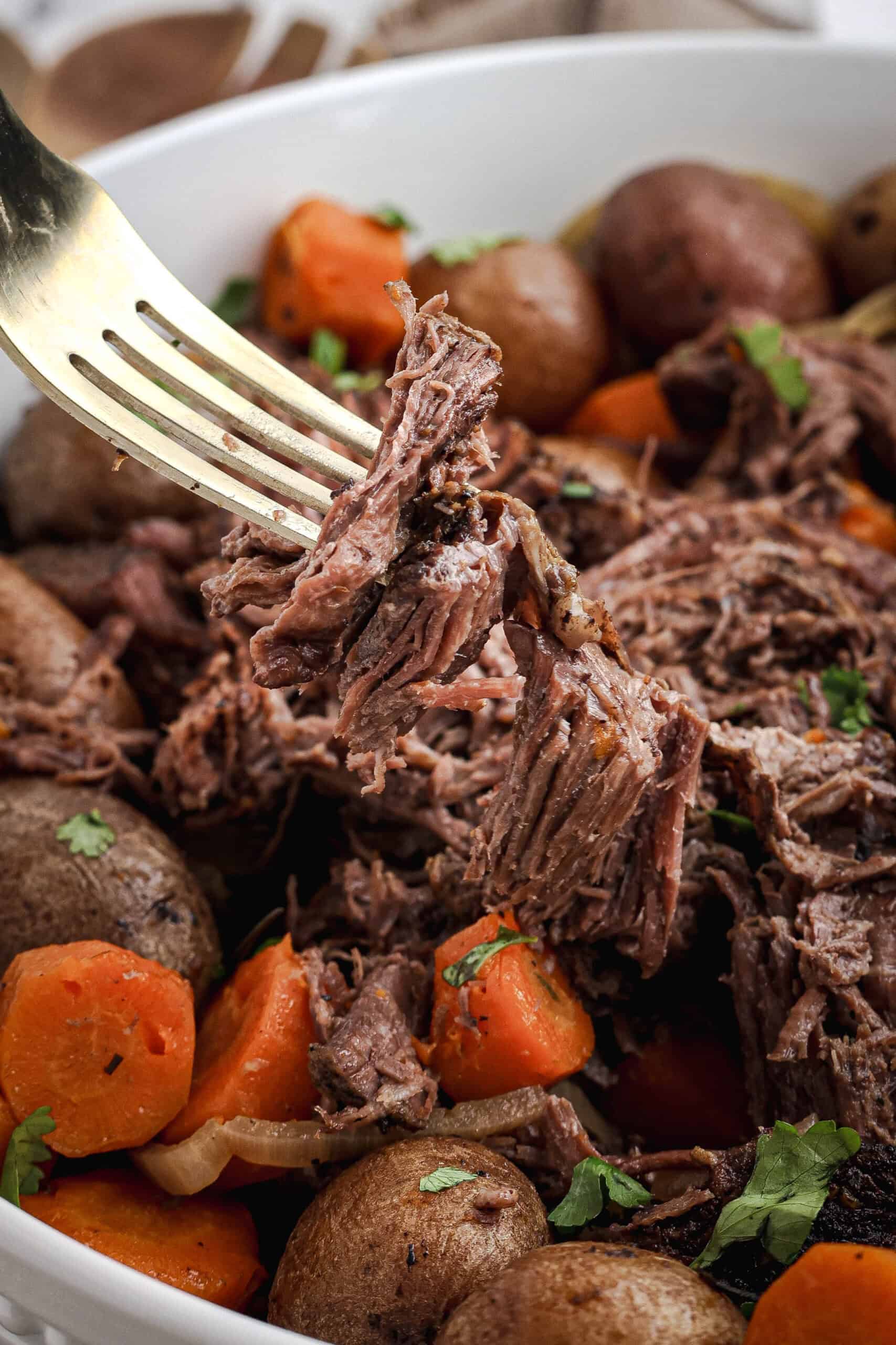 Close-up of a fork pulling apart tender pot roast with carrots and potatoes in a white serving bowl.