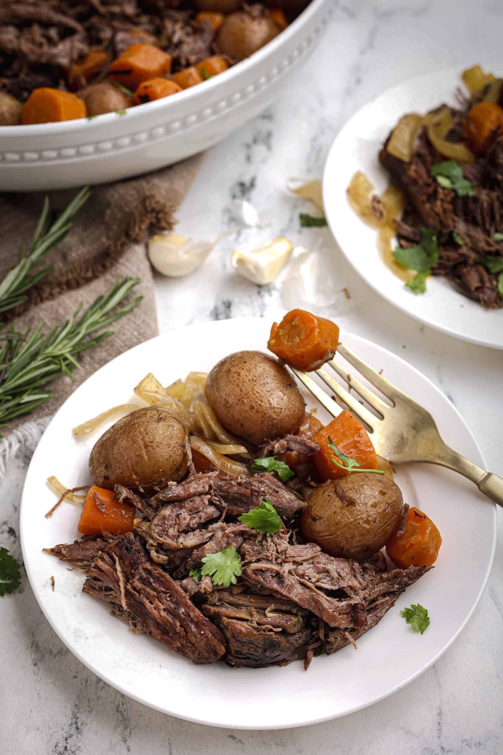 Serving plates of slow cooker pot roast with carrots and red potatoes on a white marble surface, garnished with herbs.