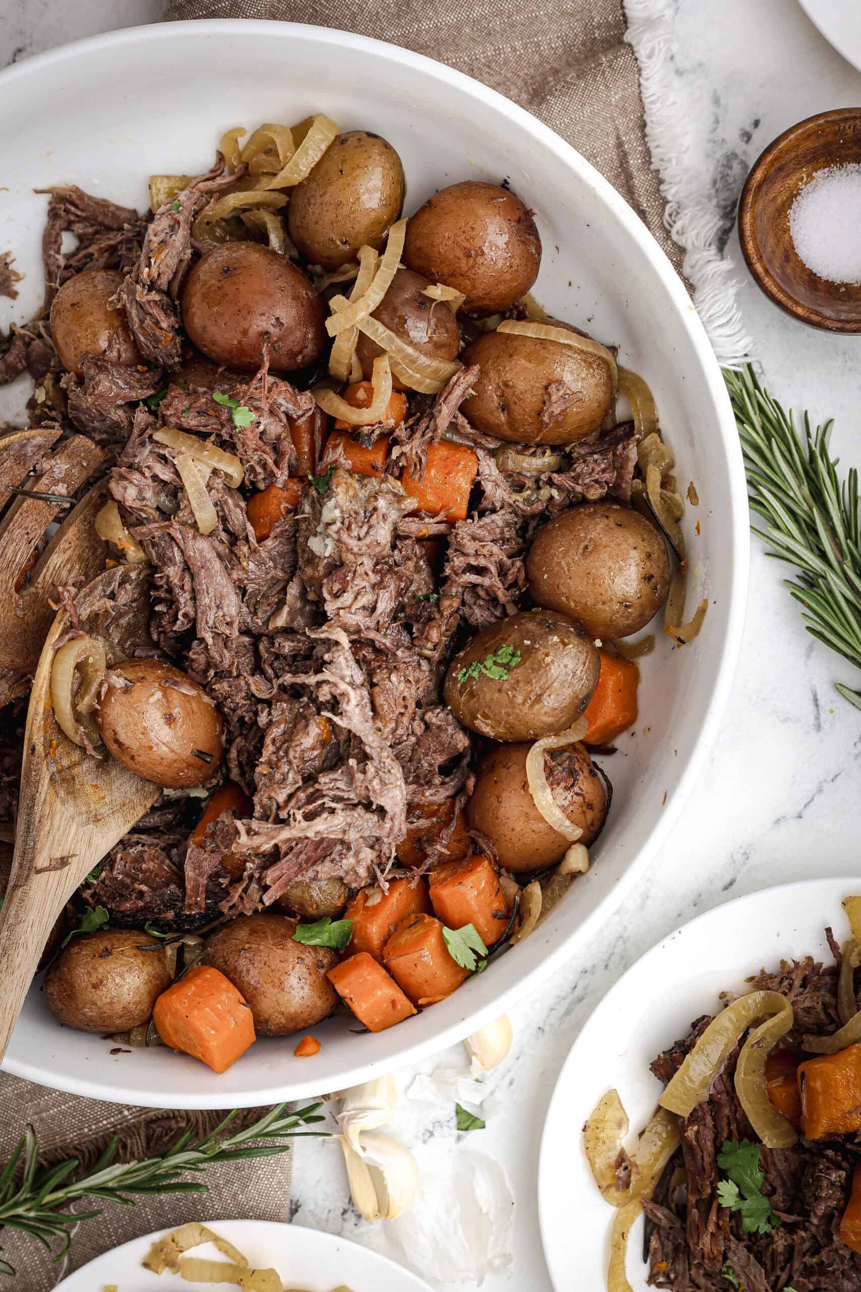 Shredded slow cooker pot roast served in a large white bowl with carrots, red potatoes, and onions, garnished with fresh herbs.