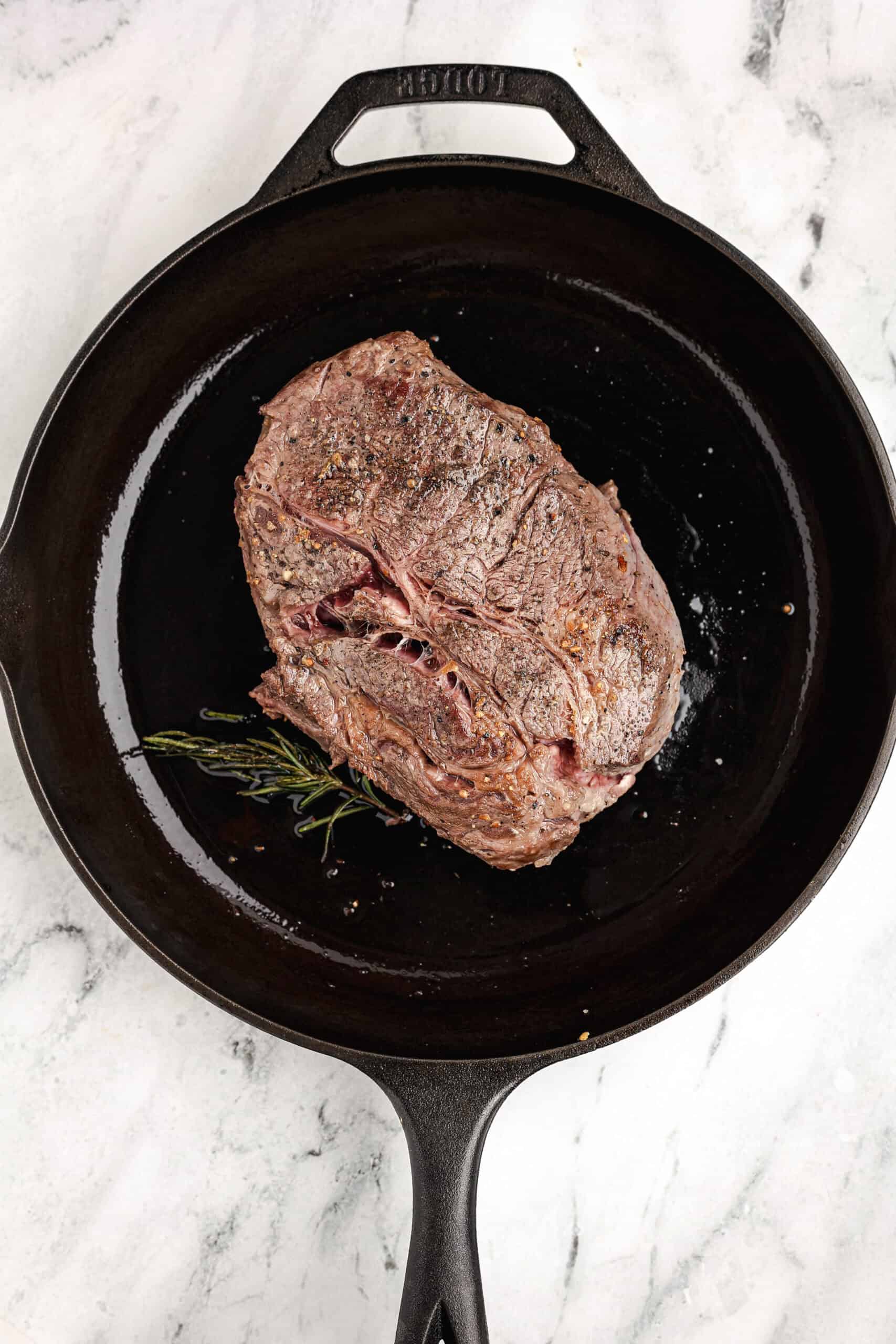 Seared chuck roast with herbs in a black cast iron skillet, showing golden-brown crust before adding to the slow cooker.