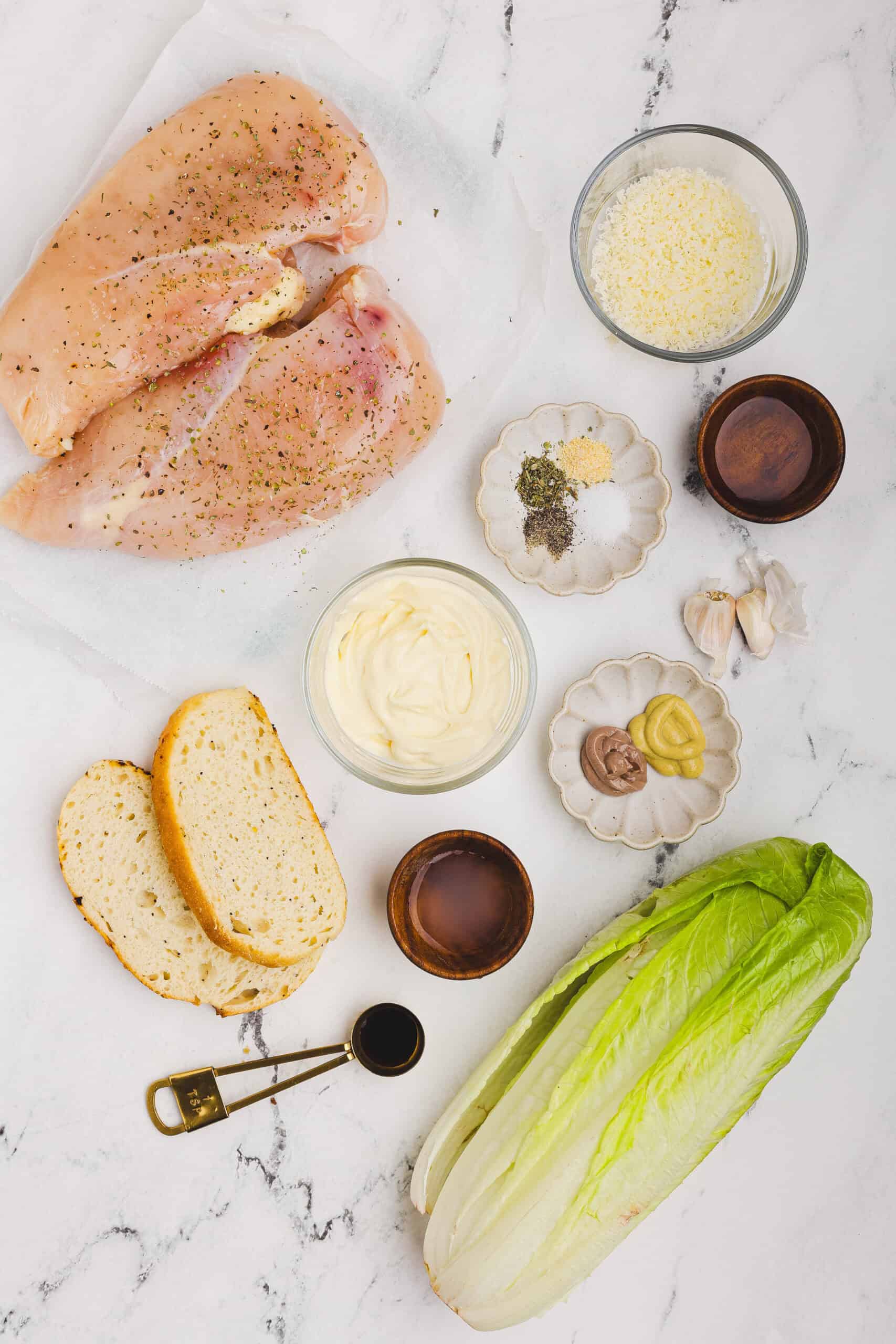 Caesar salad ingredients including raw chicken breasts, romaine lettuce, Parmesan cheese, garlic, anchovy paste, Dijon mustard, bread slices, and seasonings arranged on a marble surface