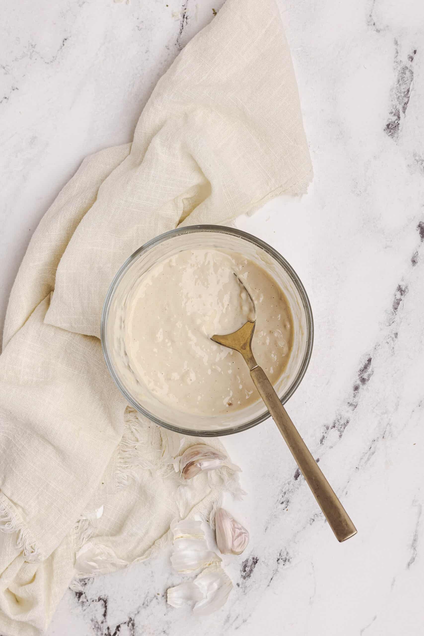 Homemade Caesar dressing in a glass bowl with a spoon, garlic cloves, and linen cloth on a marble surface