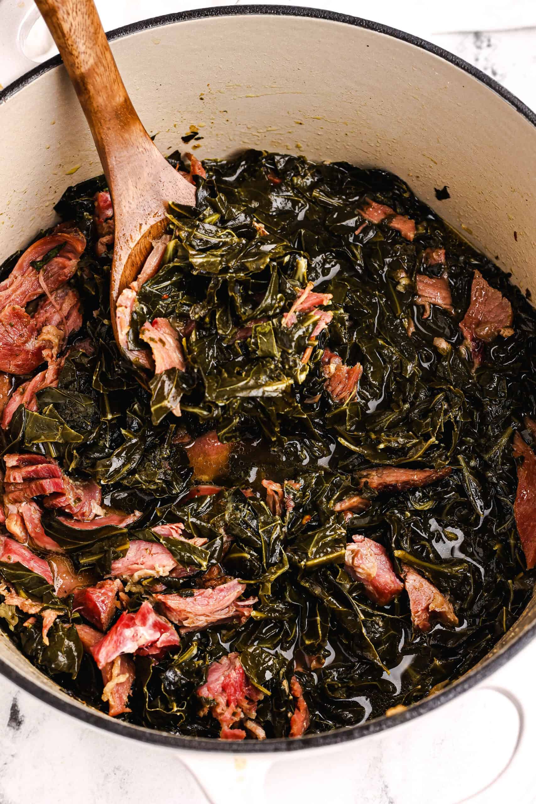 Southern collard greens cooked with ham bone in a Dutch oven, stirred with a wooden spoon.