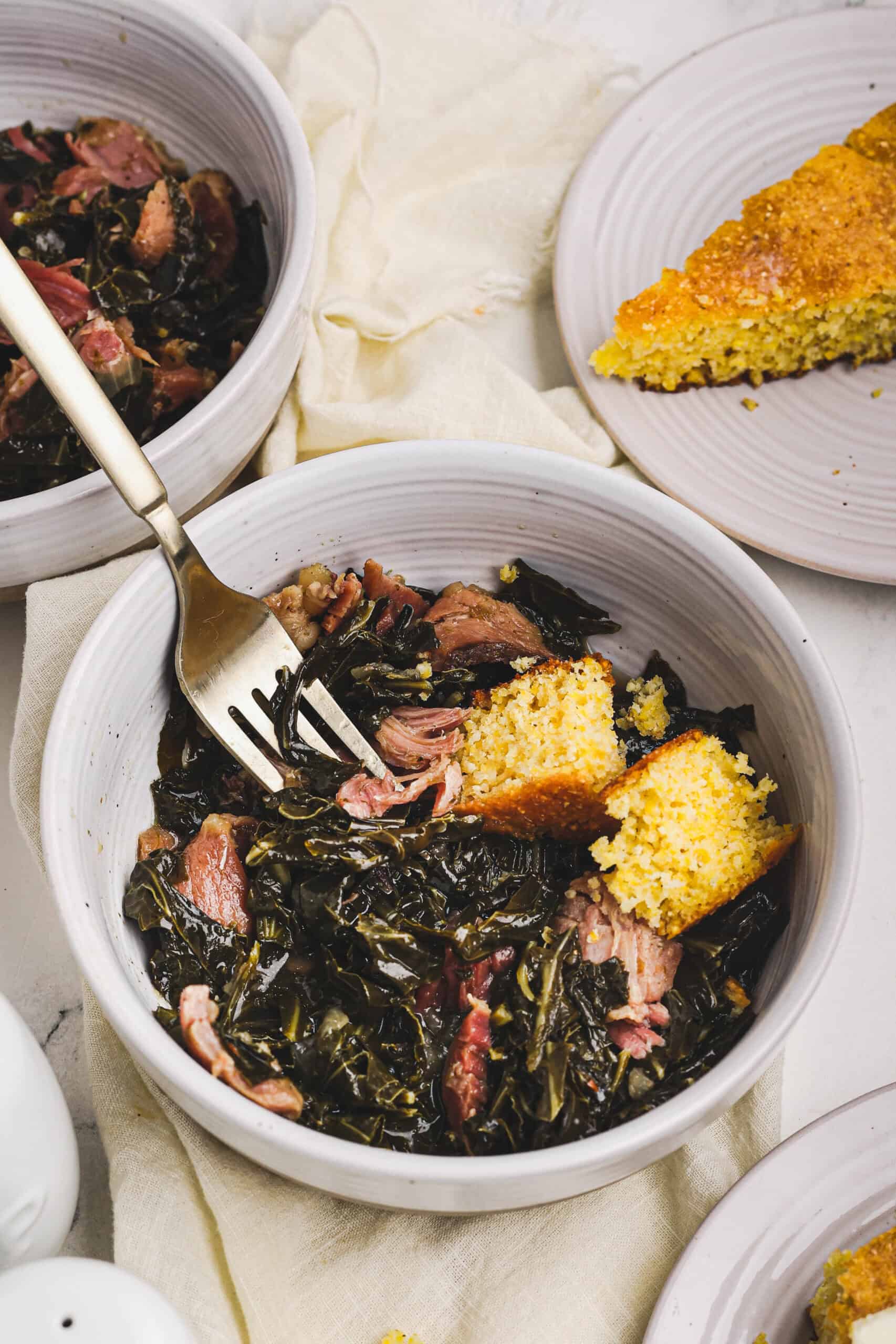 A bowl of collard greens and ham with pieces of corn bread in the bowl and to the side.