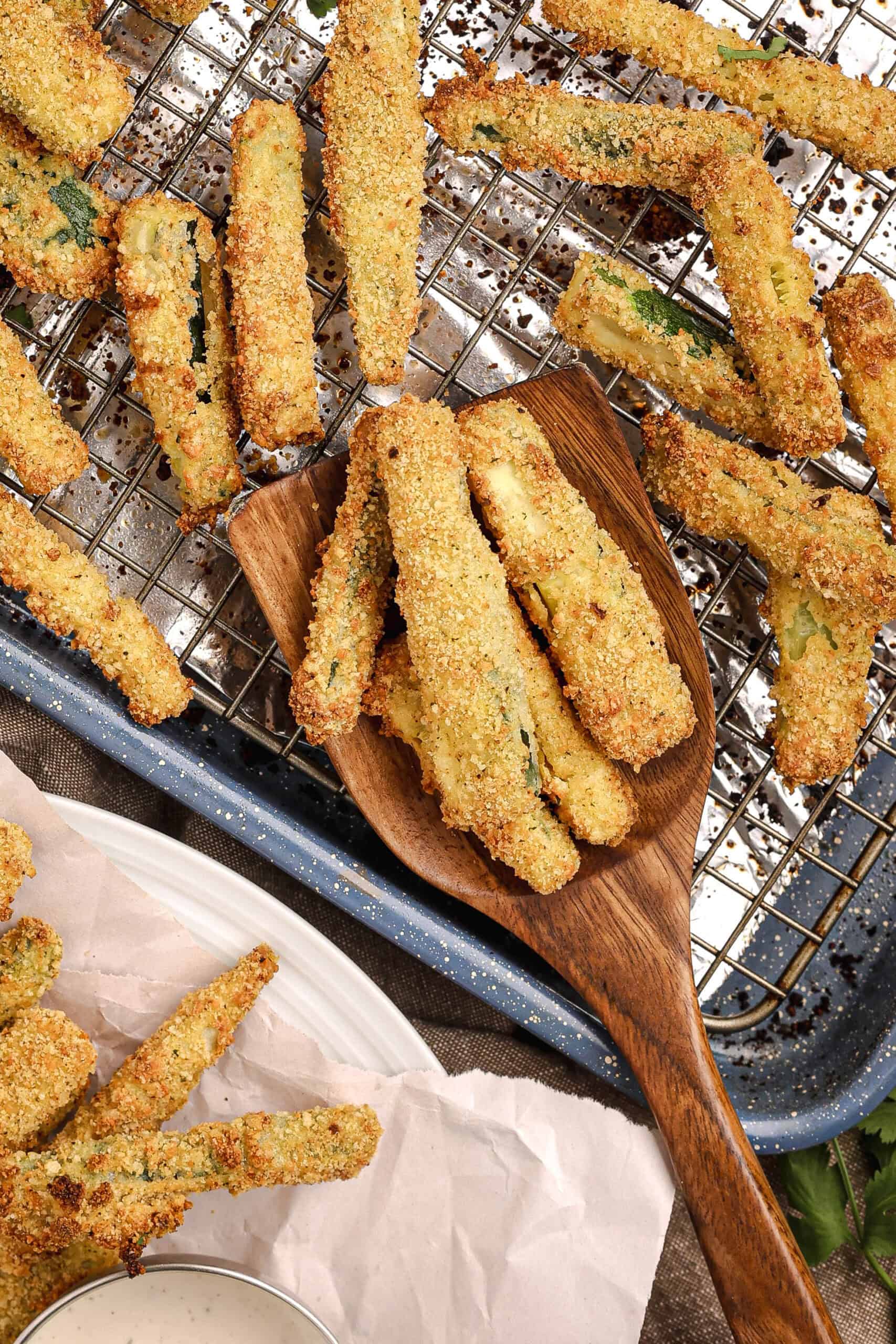 Golden baked zucchini fries resting on a wire rack with a wooden spoon holding several fries, highlighting their crisp Parmesan breadcrumb coating.