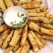 Plate of crispy baked zucchini fries served with a bowl of creamy dipping sauce, garnished with fresh herbs.