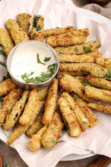 Plate of crispy baked zucchini fries served with a bowl of creamy dipping sauce, garnished with fresh herbs.