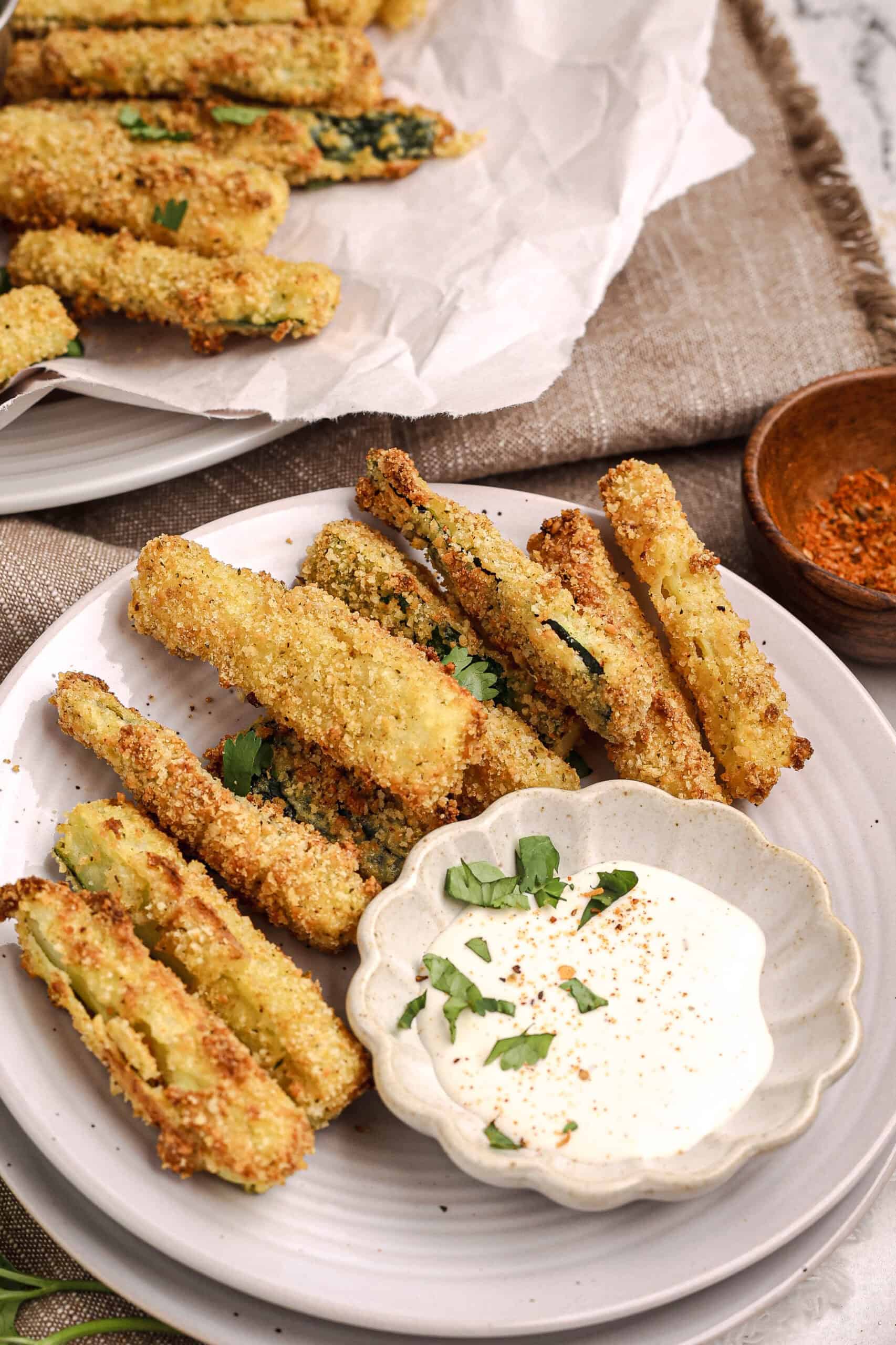 Plate of crispy baked zucchini fries served with a bowl of creamy dipping sauce, garnished with fresh herbs.