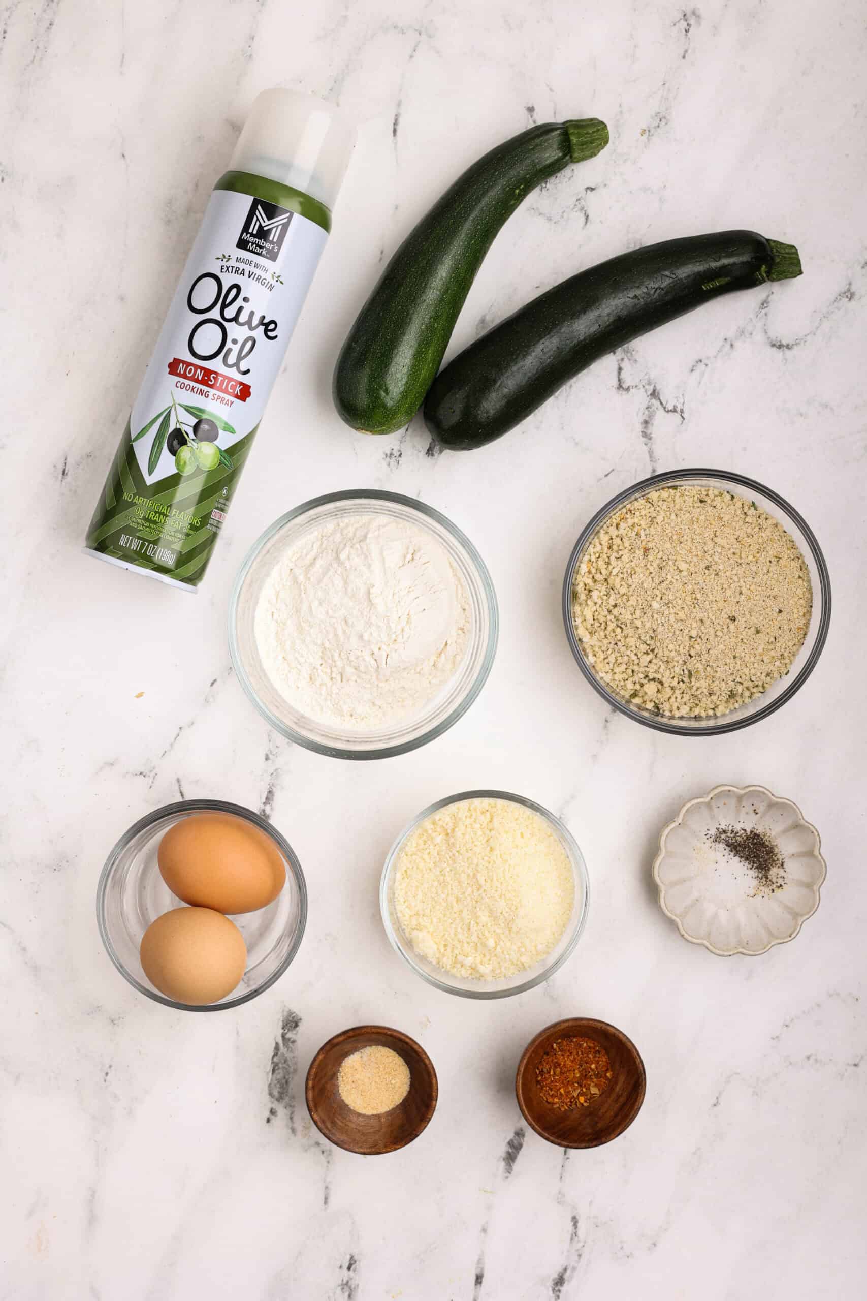 Ingredients for baked zucchini fries including fresh zucchini, eggs, flour, breadcrumbs, Parmesan cheese, Cajun seasoning, garlic powder, and olive oil spray.