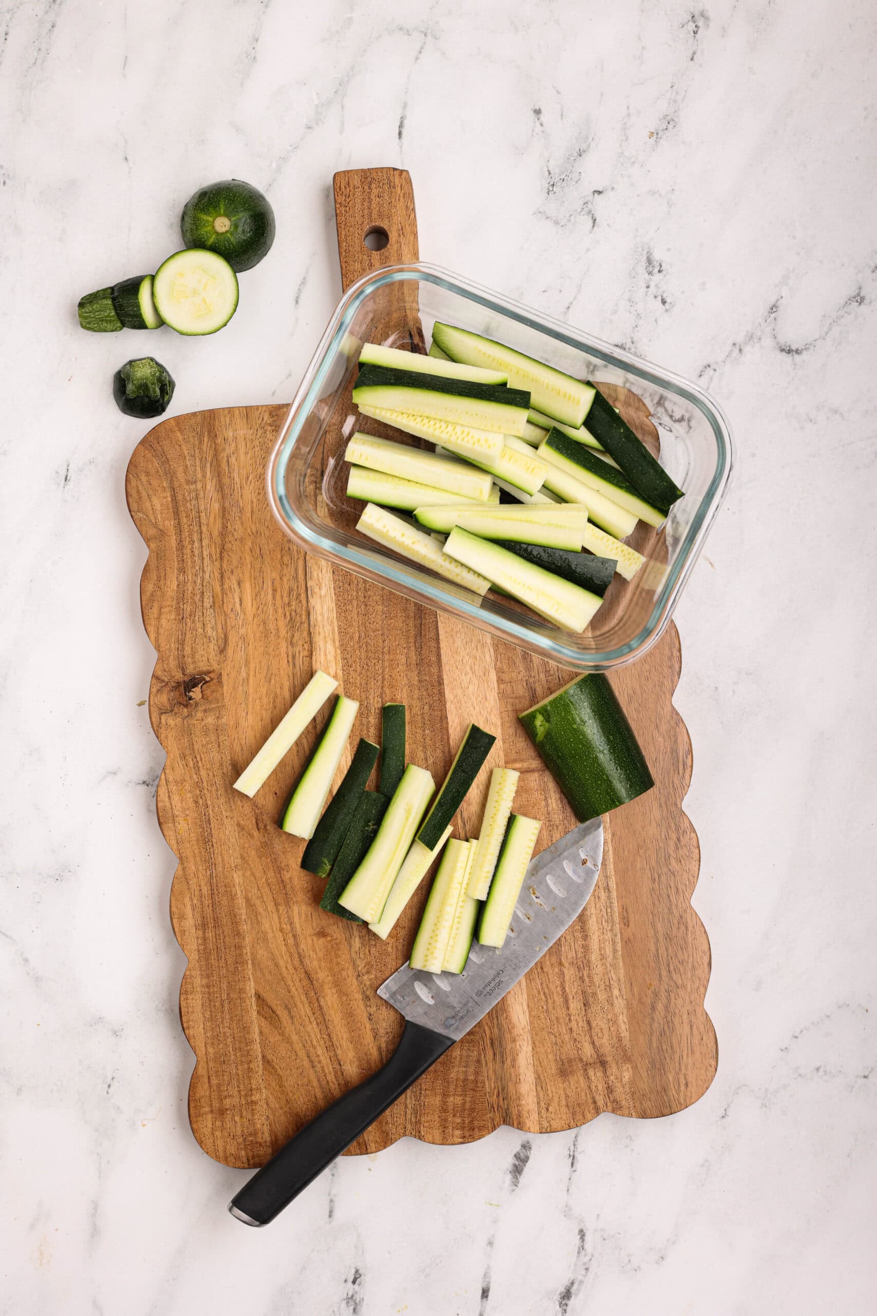 Raw zucchini cut into fry-shaped sticks arranged on a wooden cutting board with a chef’s knife.