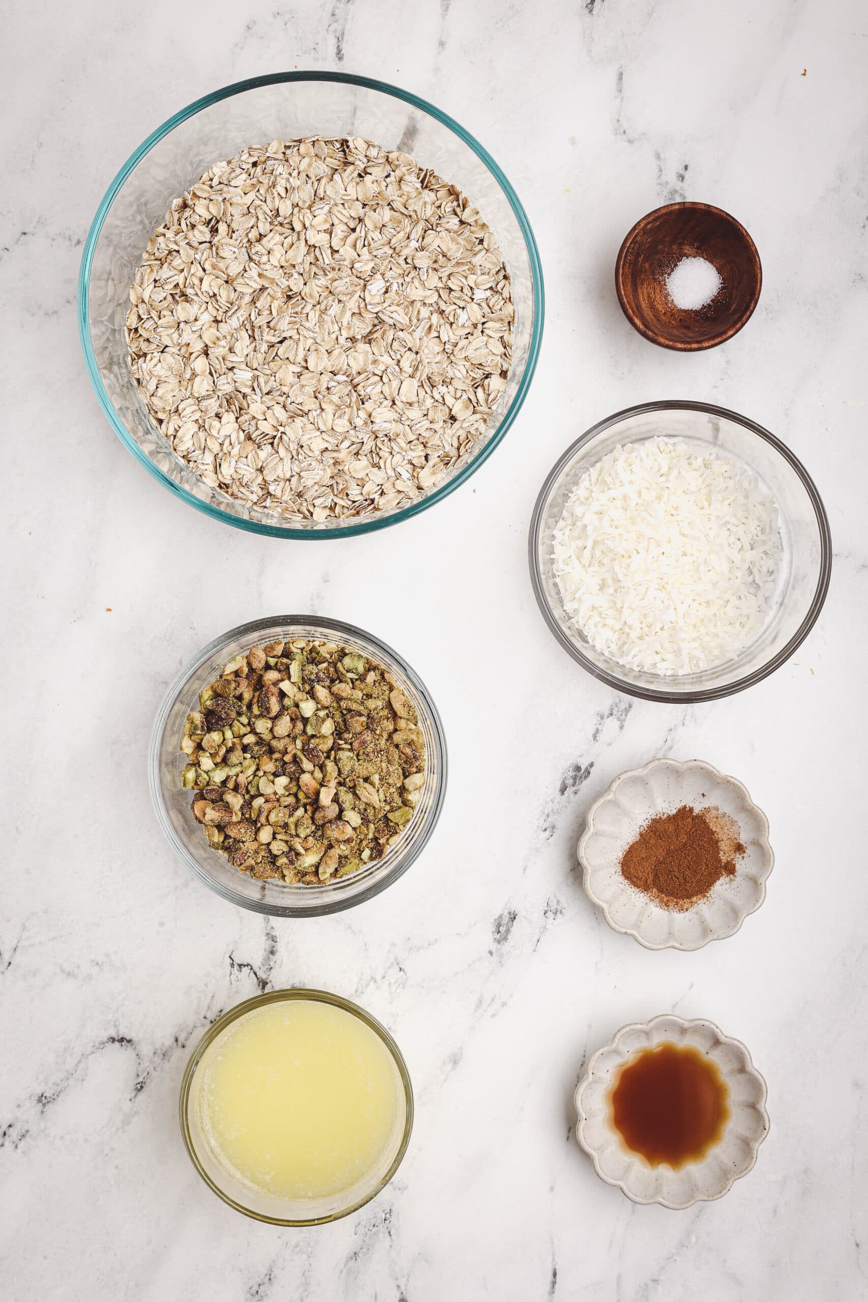 Granola ingredients including rolled oats, chopped nuts, shredded coconut, melted butter, vanilla, cinnamon, and salt on a white surface.