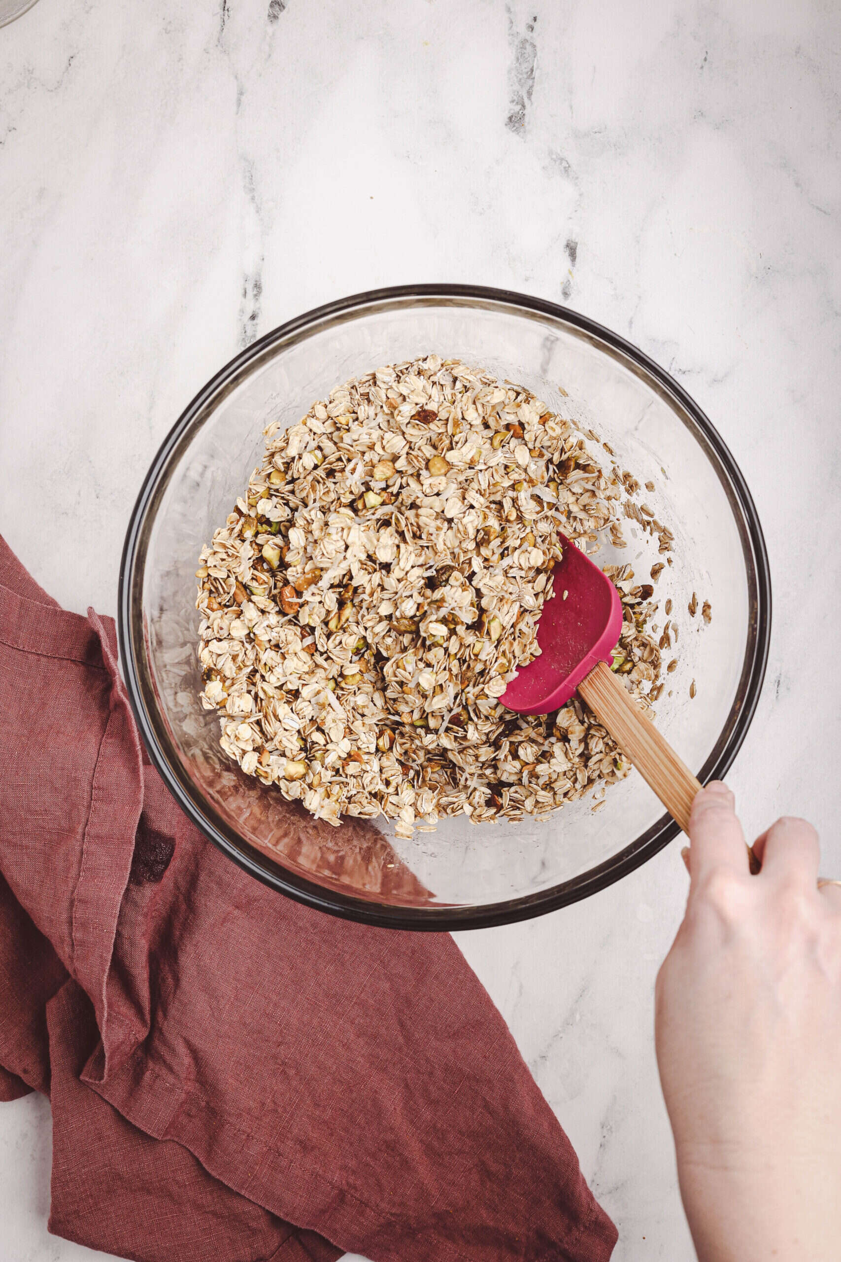 Mixing granola ingredients in a glass bowl with oats, nuts, and wet ingredients using a spatula.