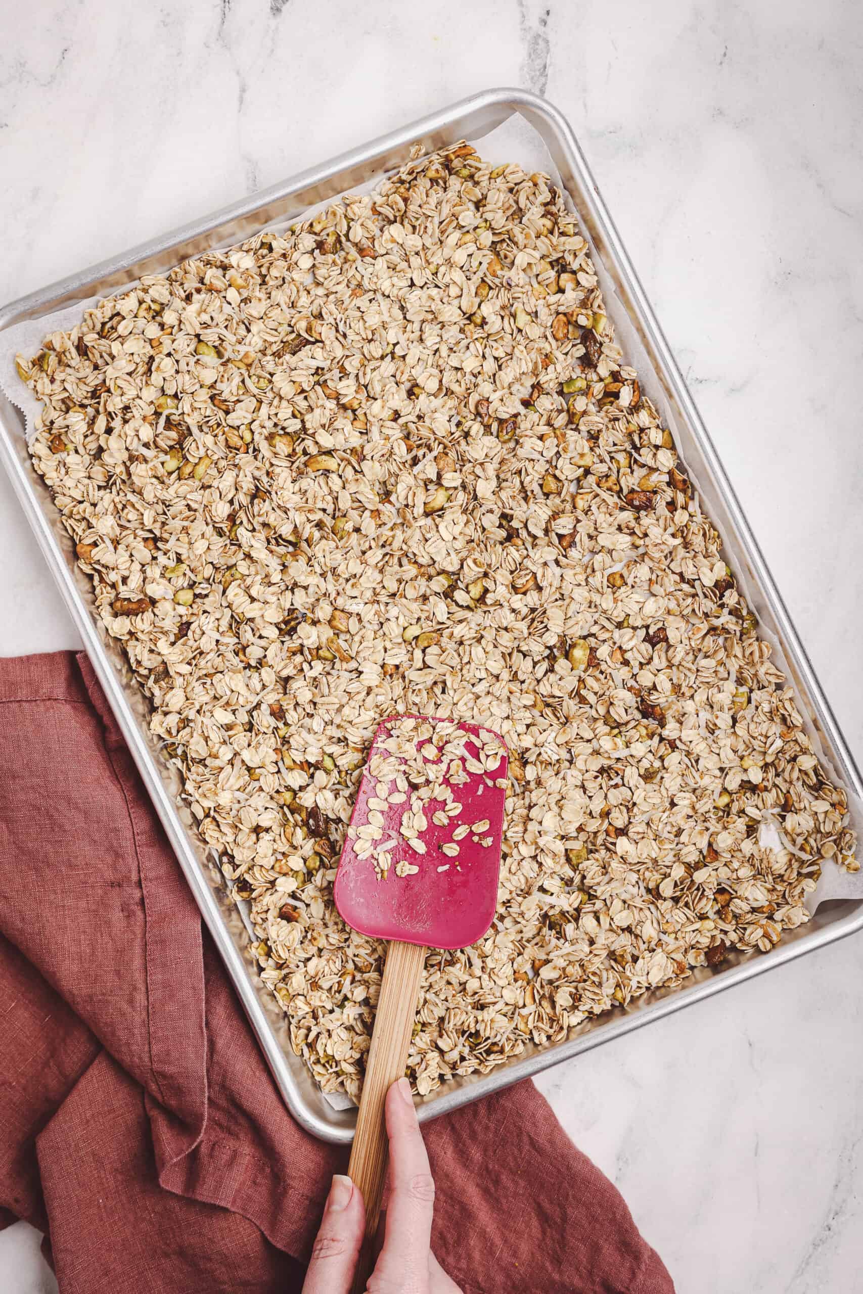Mixing granola ingredients in a glass bowl with oats, nuts, and wet ingredients using a spatula.