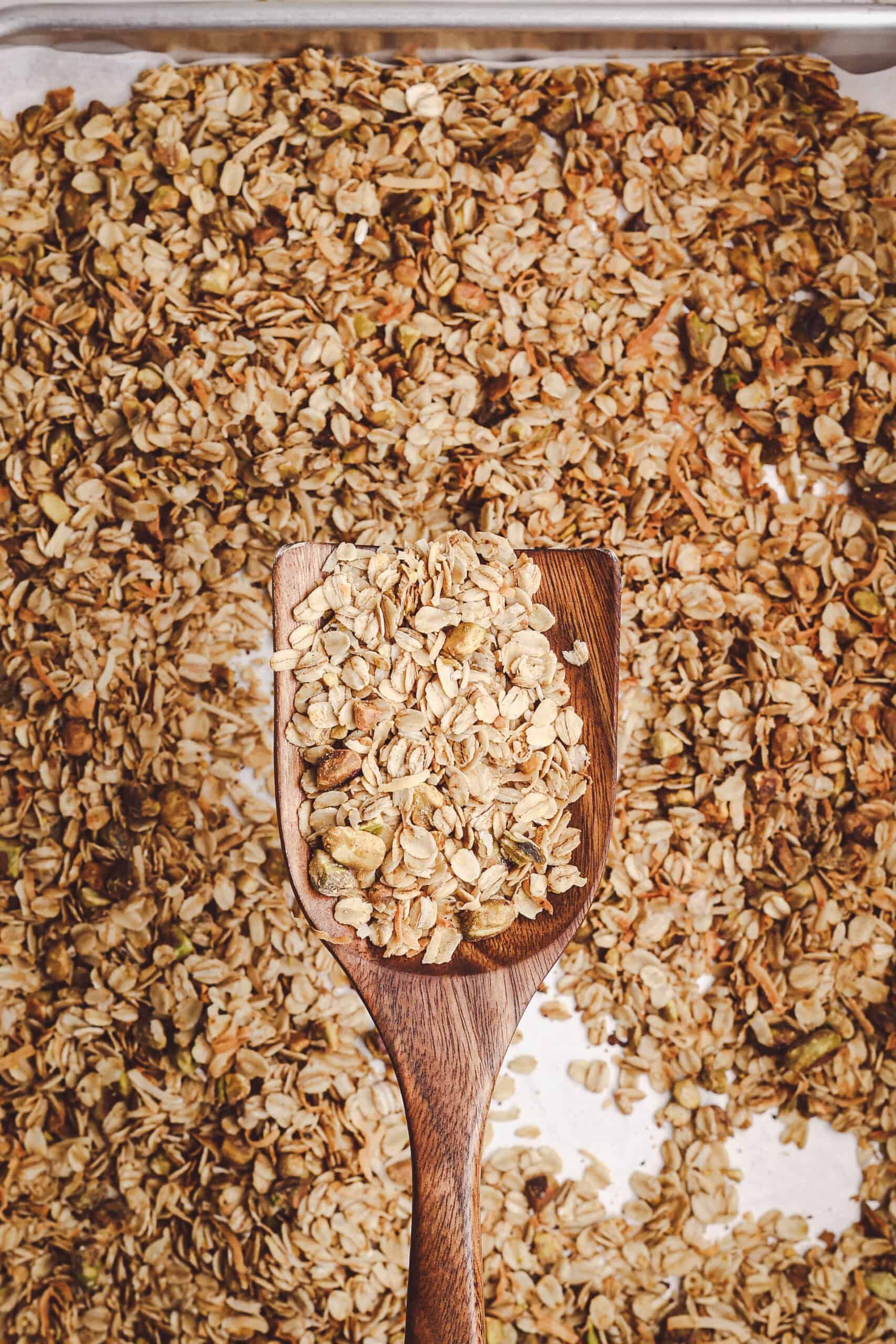 Close-up of homemade granola with oats, nuts, and coconut on a wooden spoon over a baking sheet.