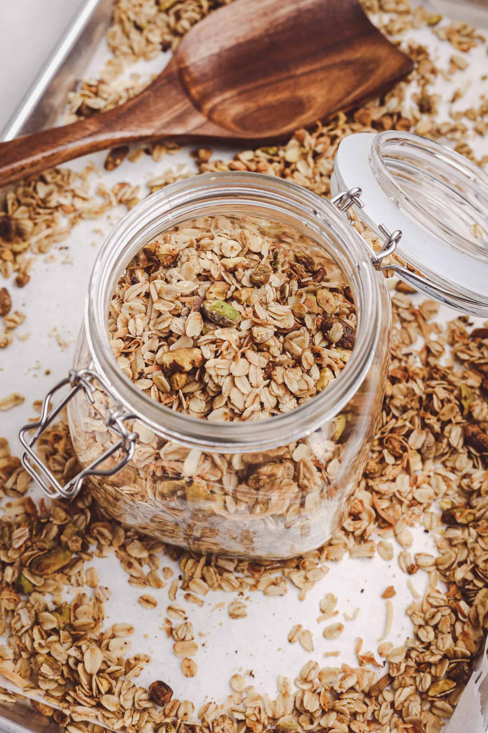Homemade granola with oats, coconut, and pistachios stored in a glass jar with a wooden spoon in the background.