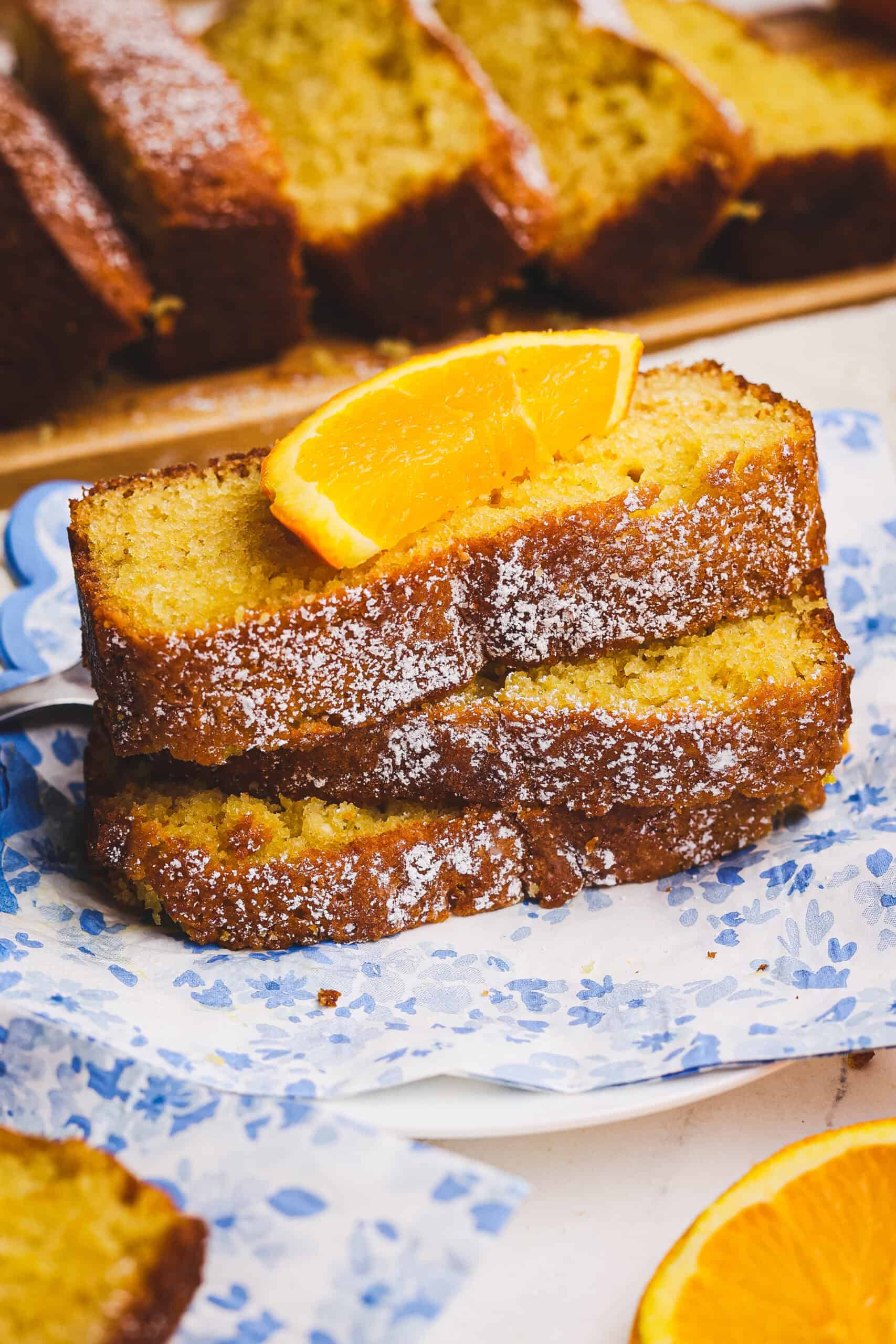 Stacked slices of orange loaf topped with powdered sugar and a fresh orange wedge on a plate with a floral napkin.