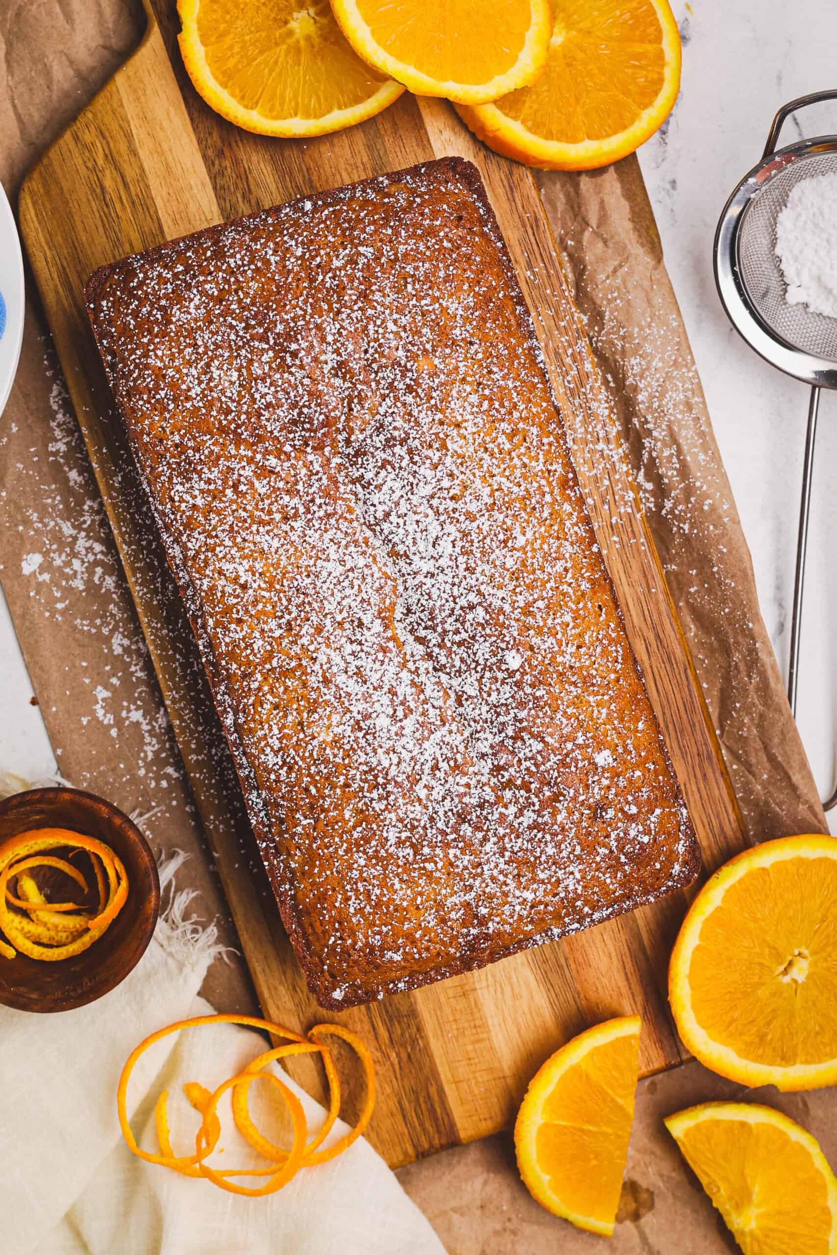 Whole orange quick loaf dusted with powdered sugar on a wooden board, surrounded by fresh orange slices and zest.