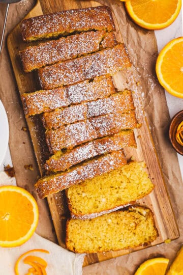 Sliced orange quick loaf dusted with powdered sugar on a wooden board, surrounded by fresh orange slices and styled with rustic parchment and linen.