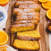 Moist orange quick loaf sliced on a wooden board, dusted with powdered sugar and surrounded by fresh orange slices, styled with a rustic linen and citrus accents.