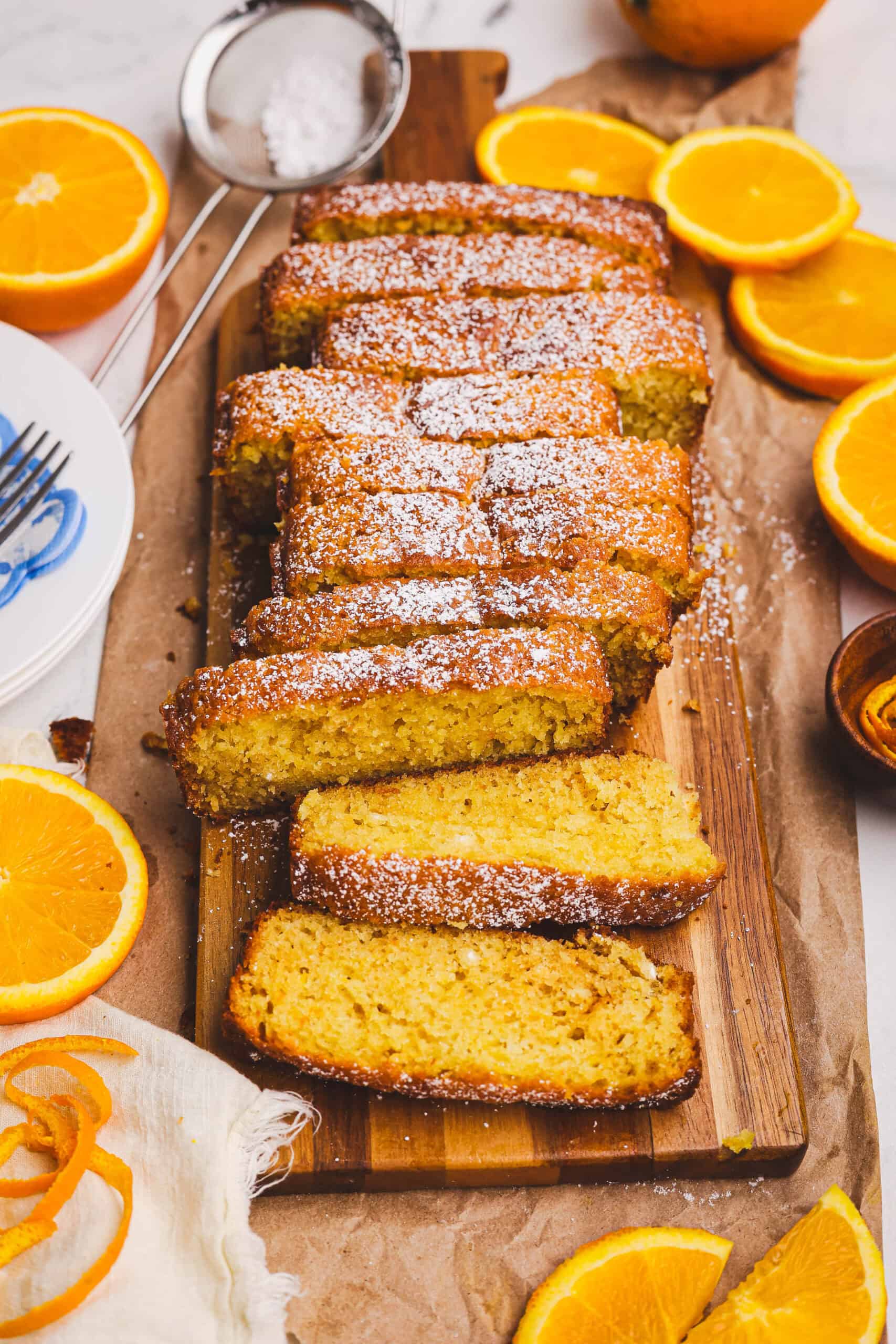 Moist orange quick loaf sliced on a wooden board, dusted with powdered sugar and surrounded by fresh orange slices, styled with a rustic linen and citrus accents.