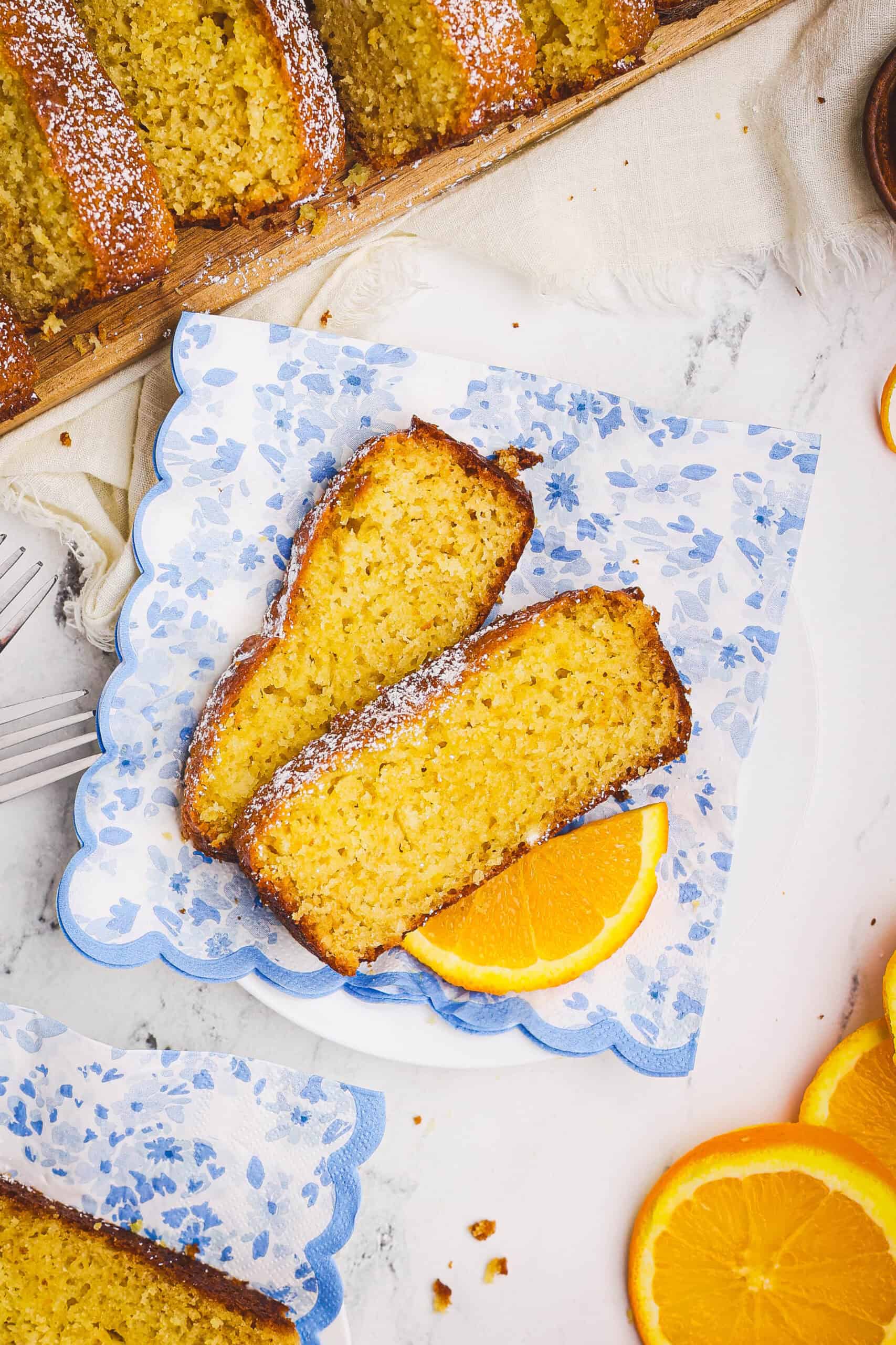 Two slices of orange quick loaf on a plate with a blue floral napkin, served with a fresh orange slice and crumbs visible.