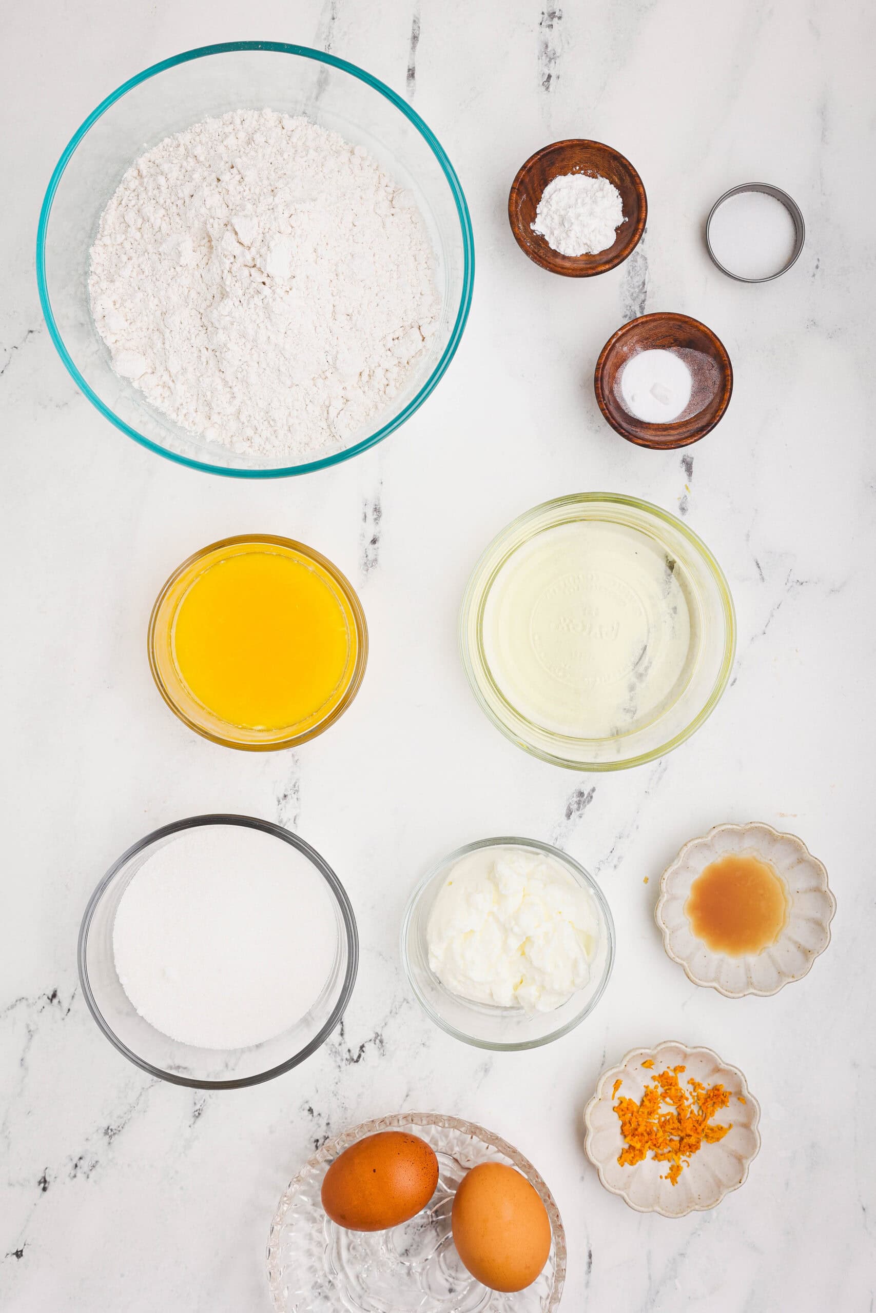 Measured ingredients for orange quick loaf including flour, sugar, eggs, orange juice, oil, yogurt, vanilla, and orange zest arranged on a marble countertop.