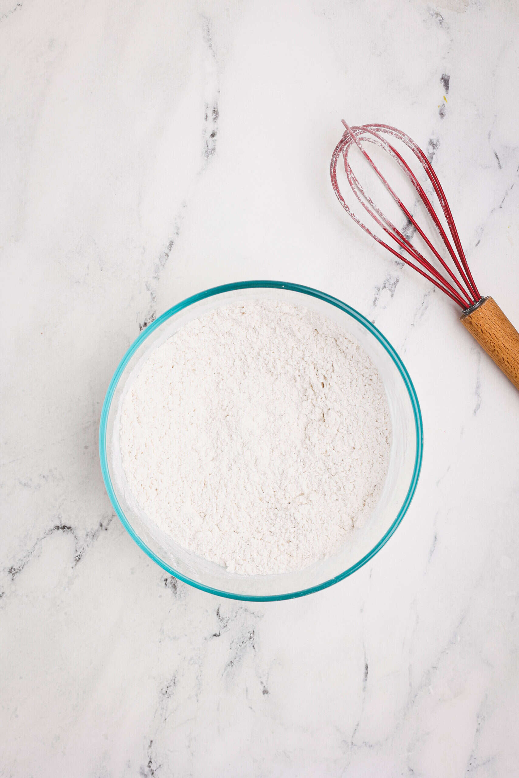 Flour in a glass mixing bowl with a whisk beside it on a marble surface.