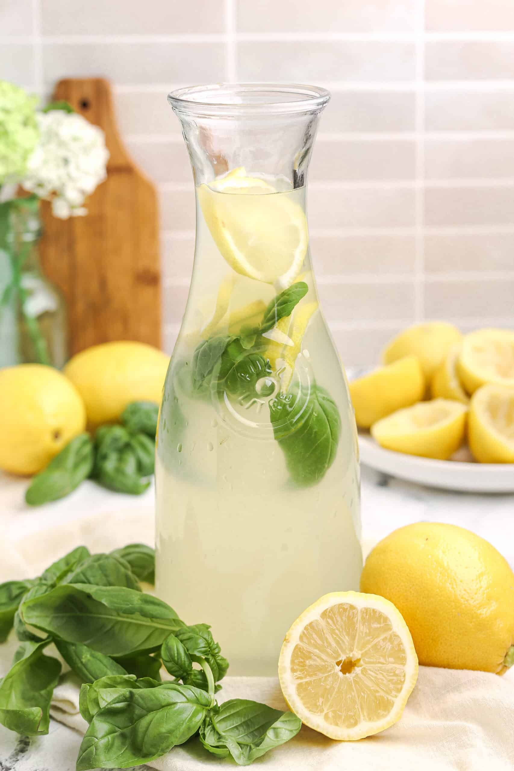 Glass carafe filled with basil lemonade, lemon slices, and fresh basil leaves on a light kitchen counter.