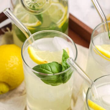 Fresh basil lemonade served over ice in clear glasses, garnished with lemon slices and basil leaves, with a glass pitcher in the background.