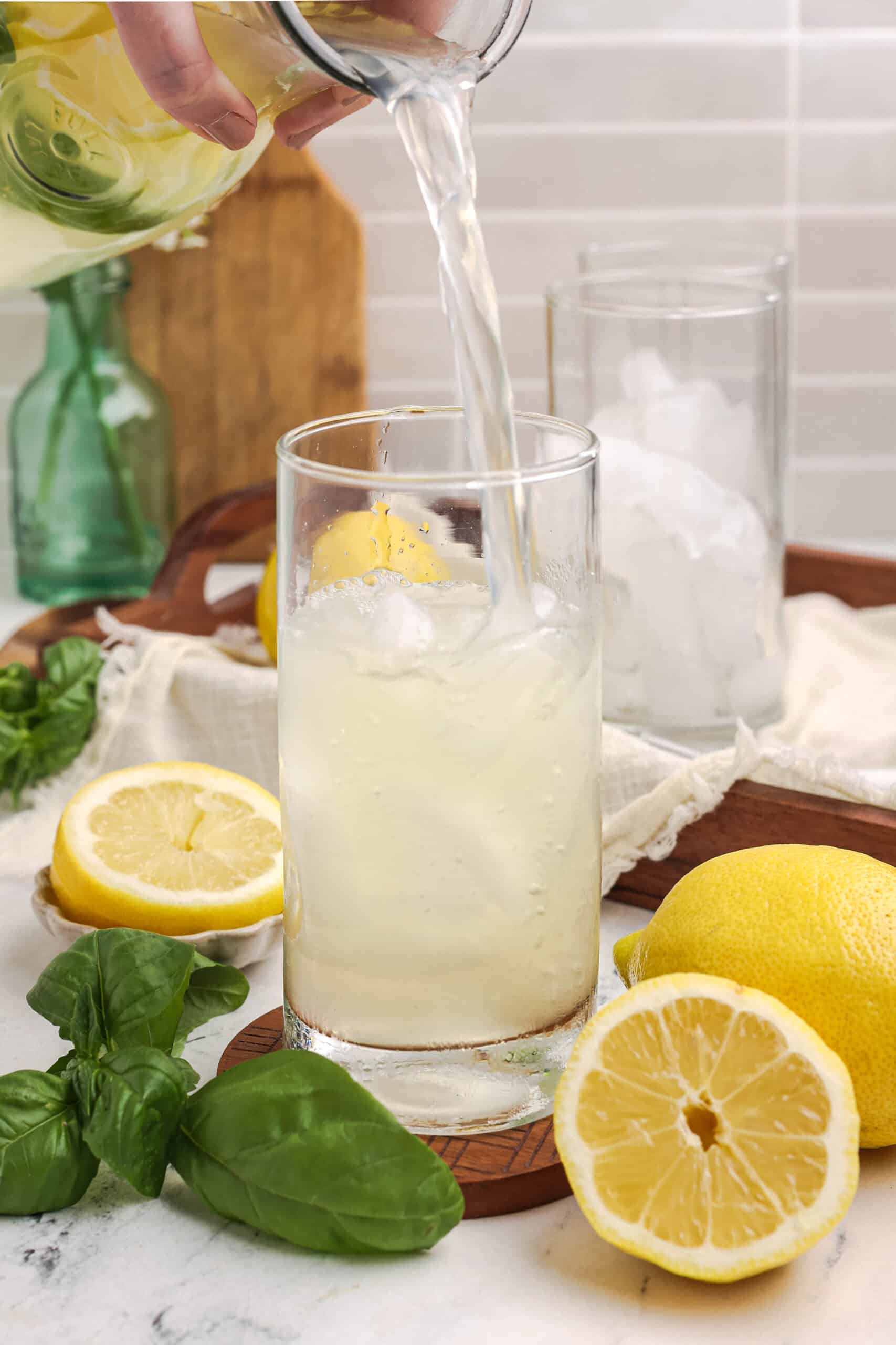 Pouring fresh basil lemonade into a glass over ice, with lemons and basil leaves styled on a kitchen counter.
