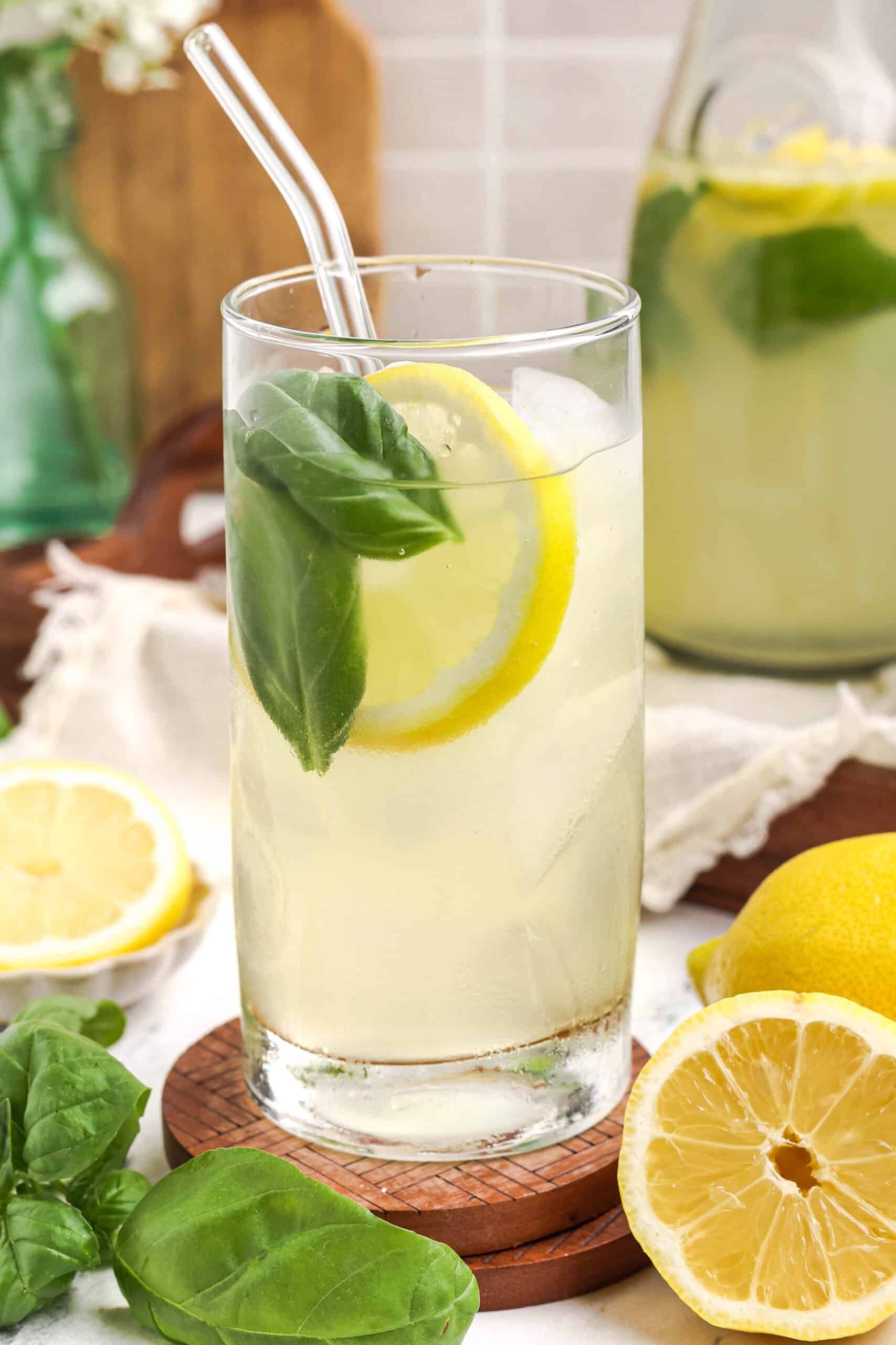 Glass of fresh basil lemonade over ice, garnished with a lemon slice and basil leaf, styled with whole lemons and a pitcher in the background.