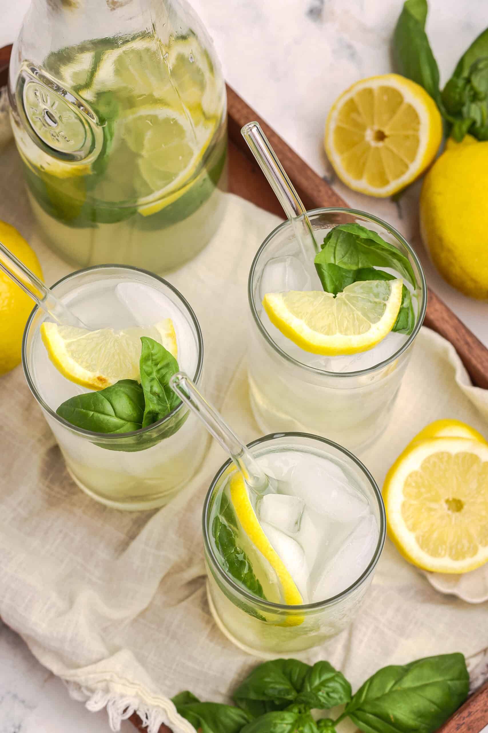 Overhead view of three glasses of basil lemonade with ice, lemon slices, and basil on a serving tray.