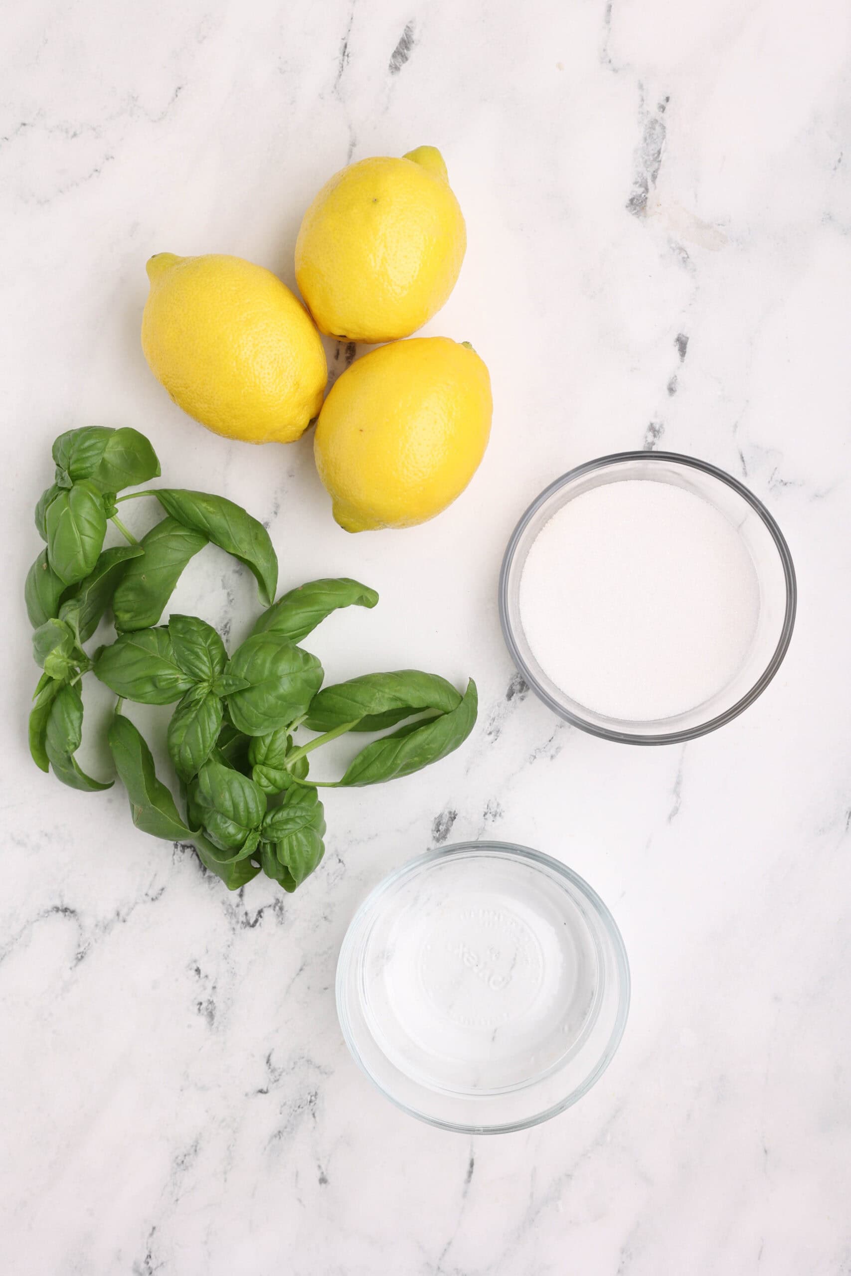 Fresh lemons, basil leaves, sugar, and water arranged on a marble surface for basil lemonade ingredients.