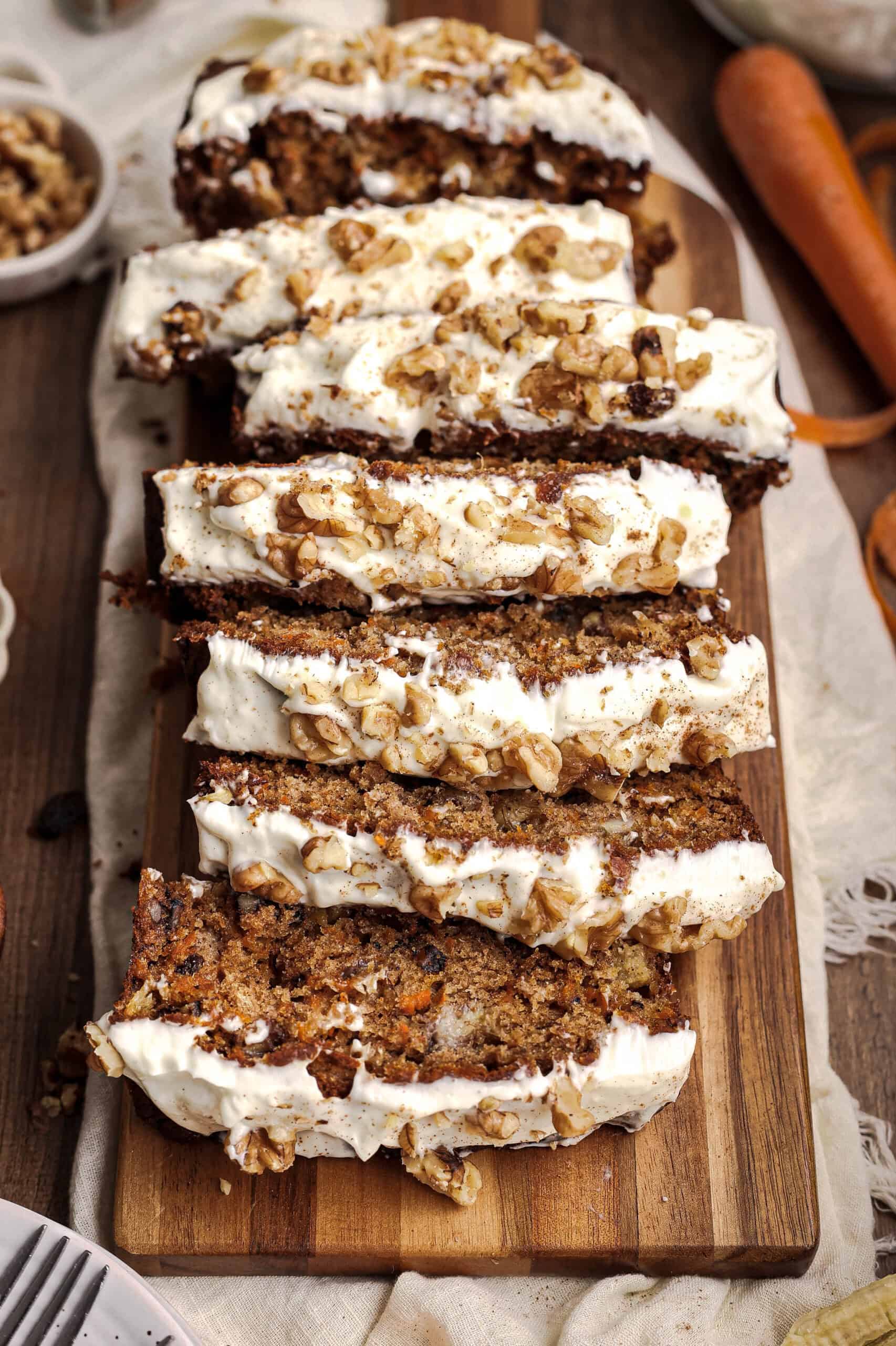 Sliced carrot cake banana bread with cream cheese frosting and chopped walnuts arranged on a wooden serving board