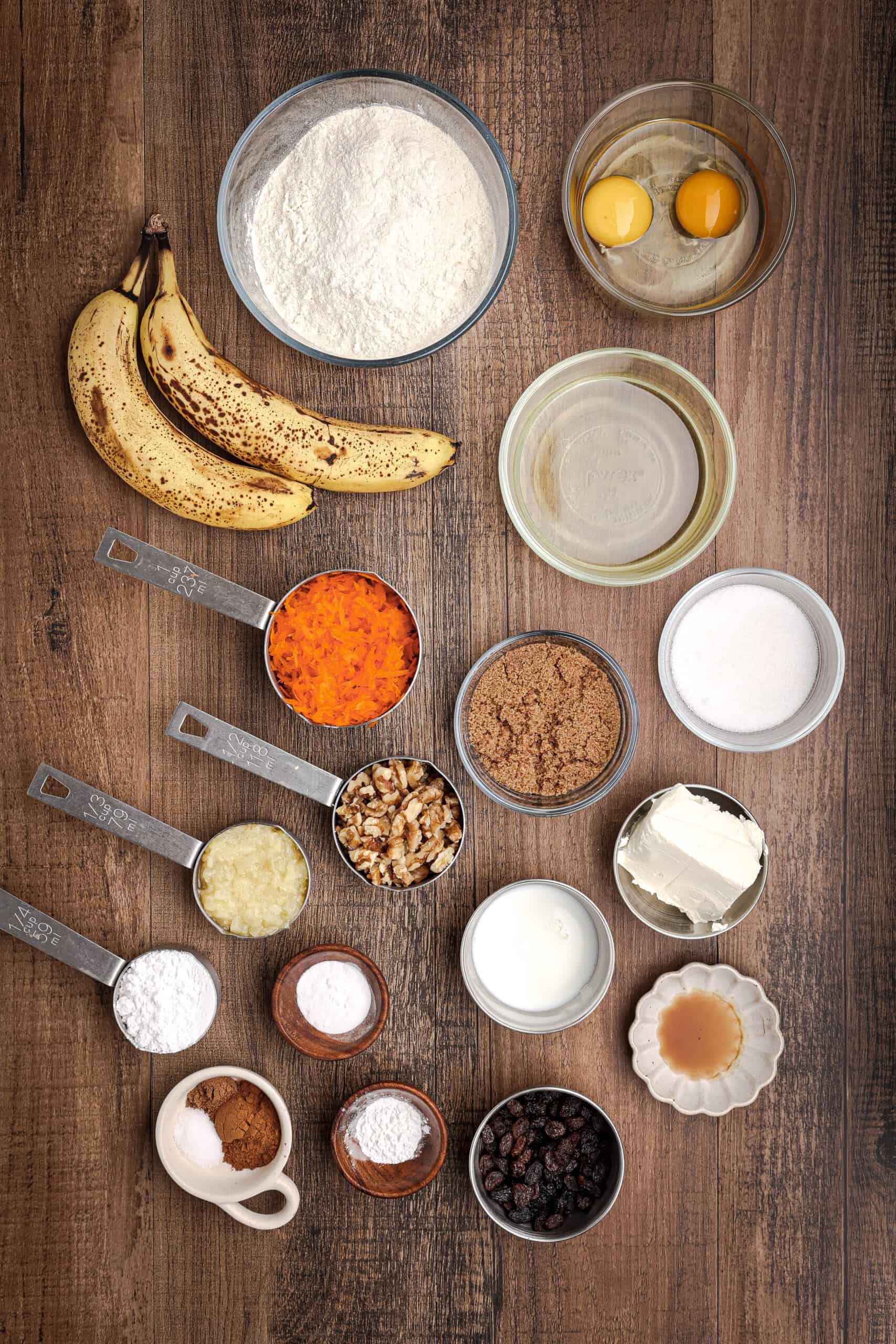 Ingredients for carrot cake banana bread including ripe bananas, flour, eggs, grated carrots, sugars, walnuts, crushed pineapple, and spices arranged on a wooden surface