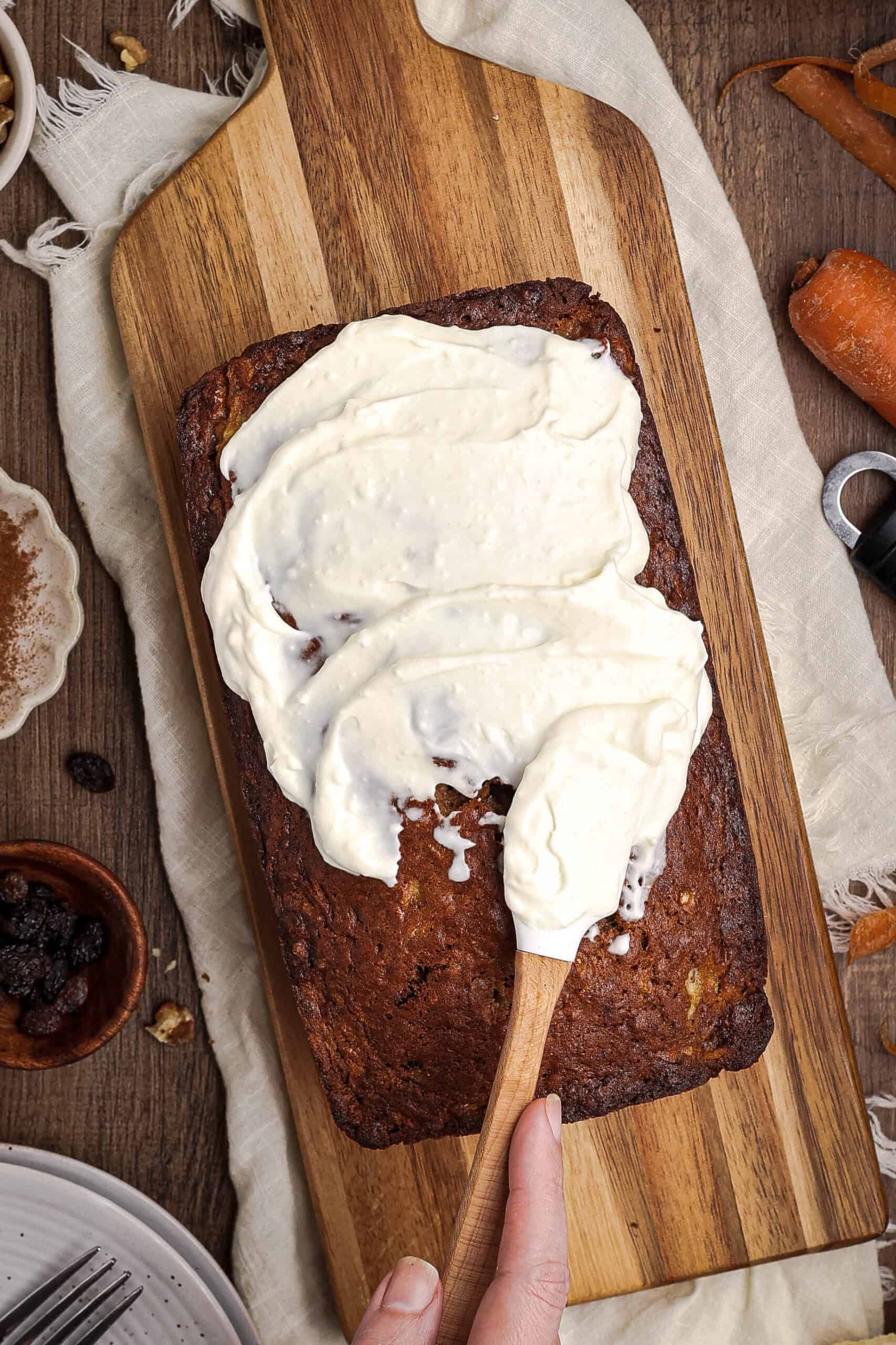 Spreading cream cheese frosting over carrot cake banana bread loaf on a wooden board